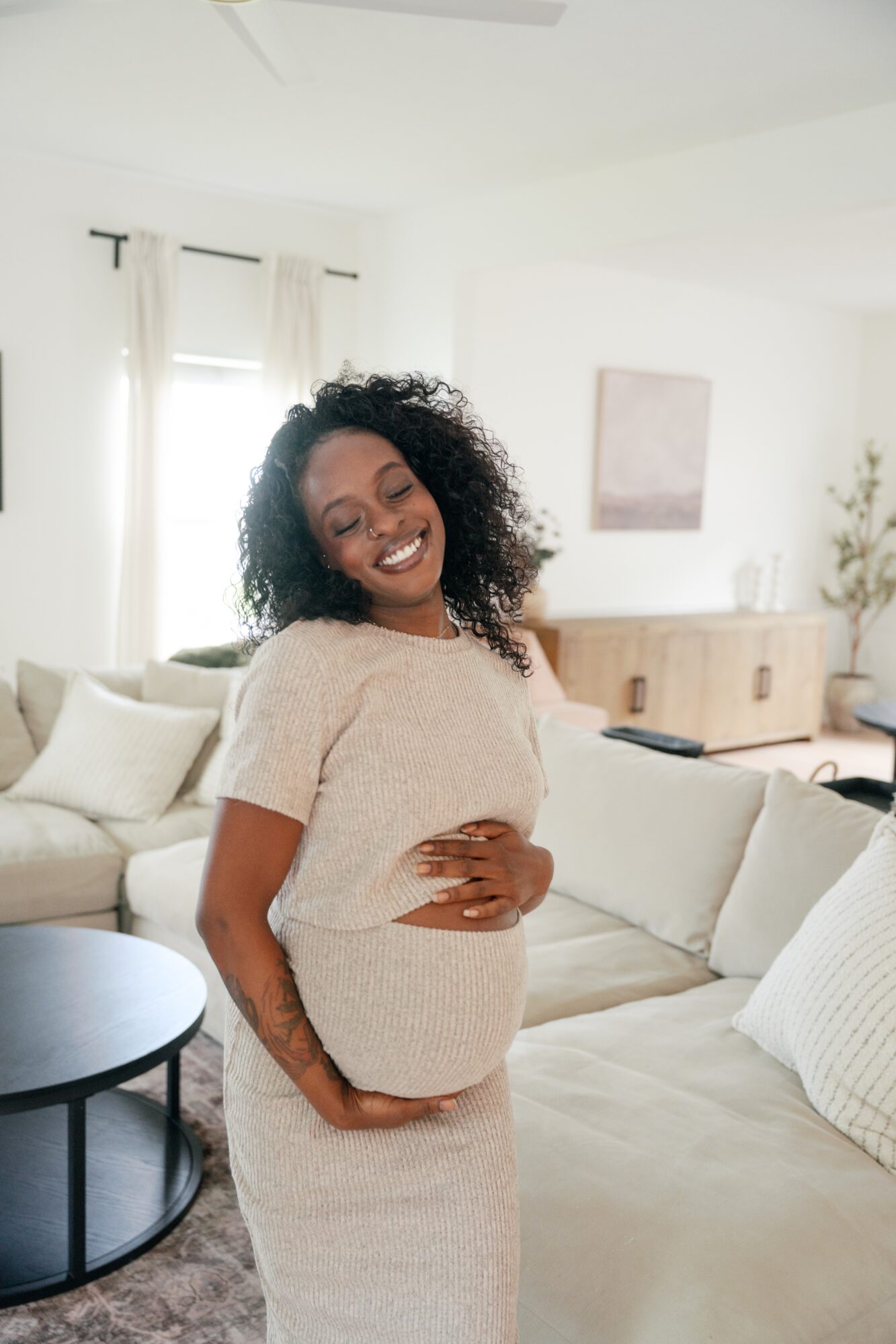 Pregnant woman with curly hair smiling and holding her belly in a bright living room with white walls and furniture.