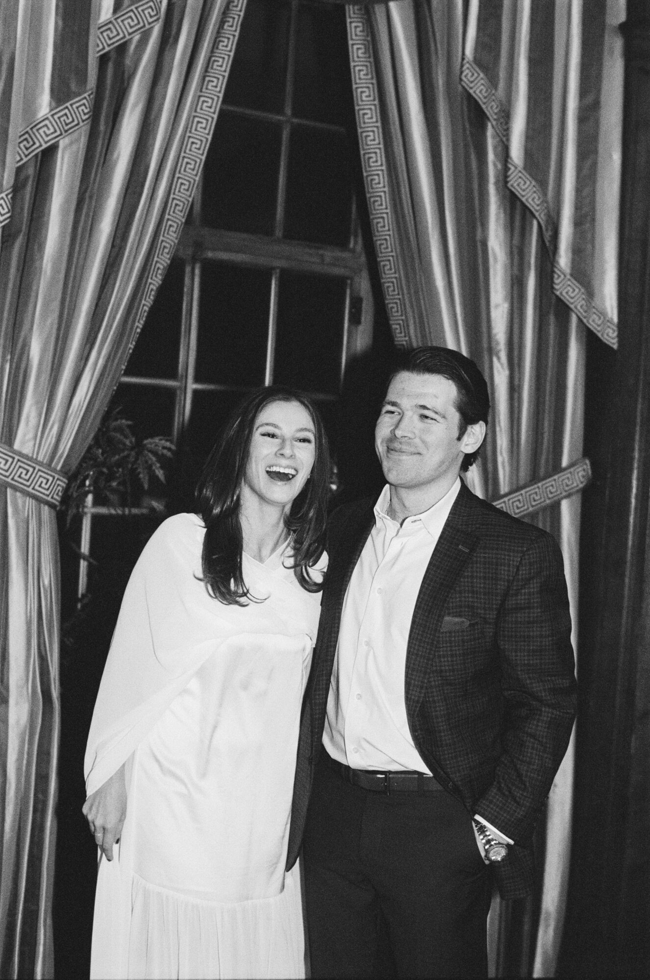 Two people, a woman and a man, standing indoors in front of a window with curtains, smiling. Black and white photo.