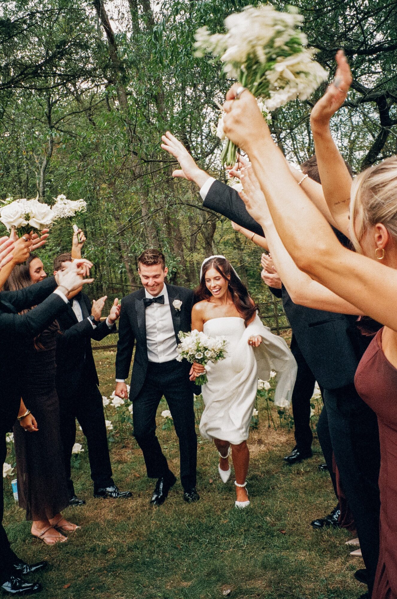 Bride and groom walking through a crowd of people holding bouquets outdoors, celebrating, with trees in the background.