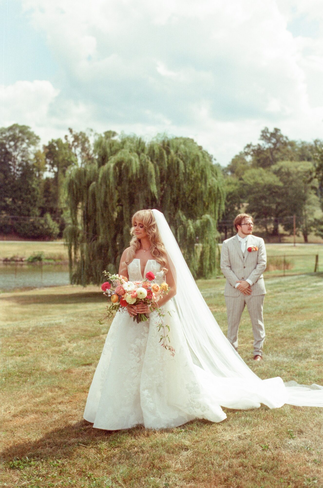 Bride in a white wedding dress holding a bouquet, standing outdoors with a groom in a suit in the background.