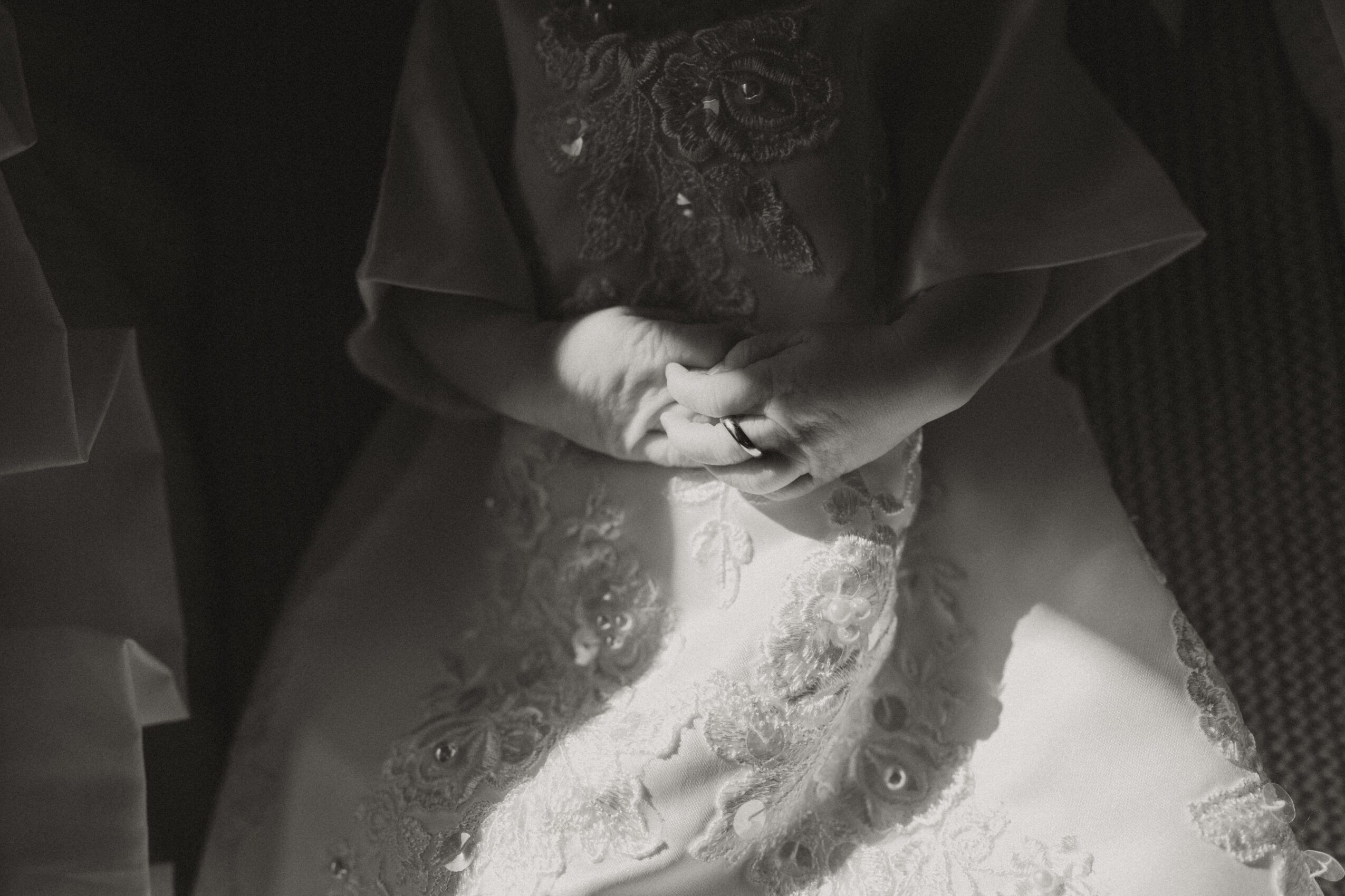 Person in a wedding dress with hands clasped, surrounded by dark clothing, in black and white.