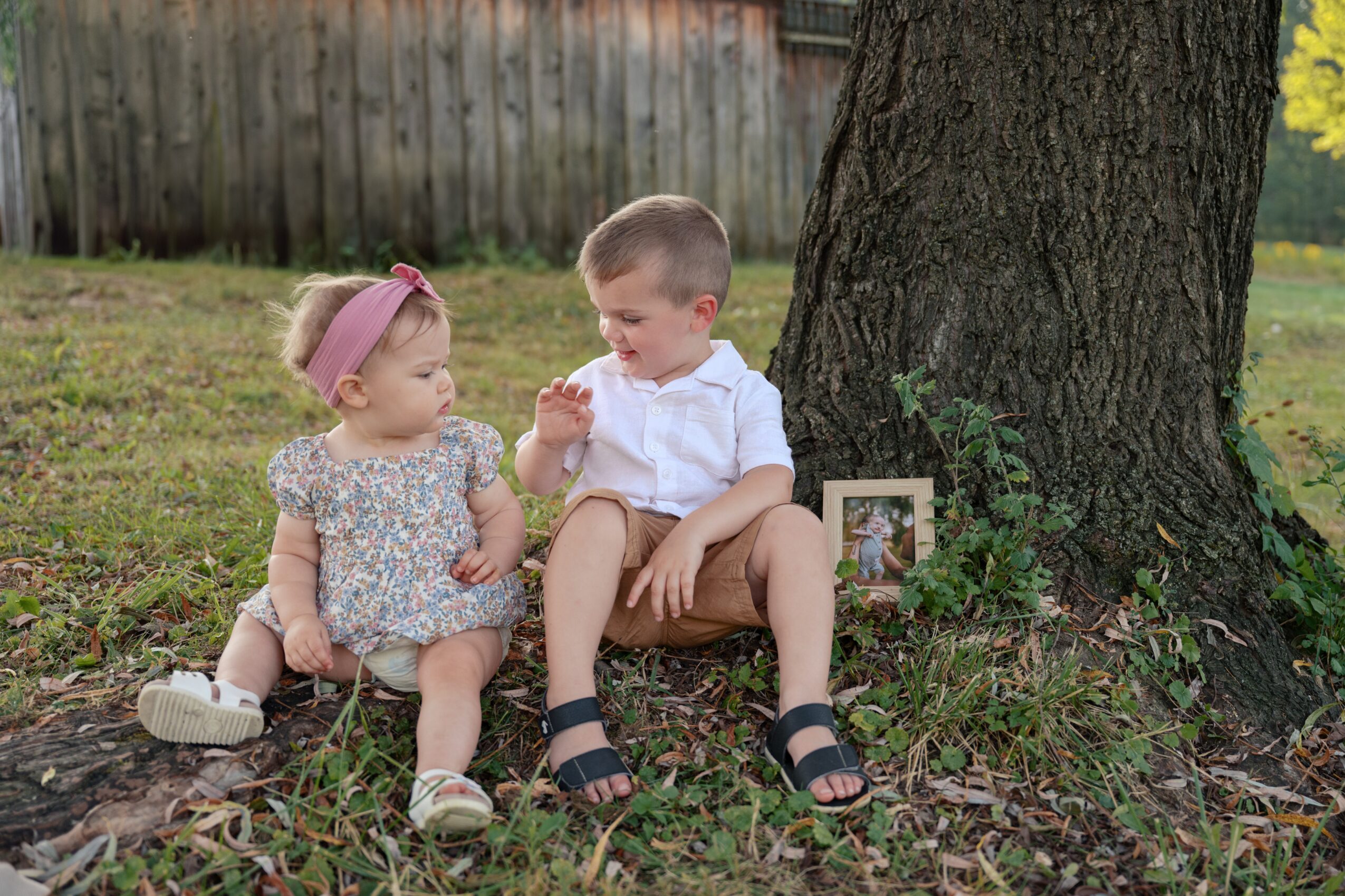 Two children sitting on grass near a tree, with a small framed photo on the ground, outdoors during daytime.