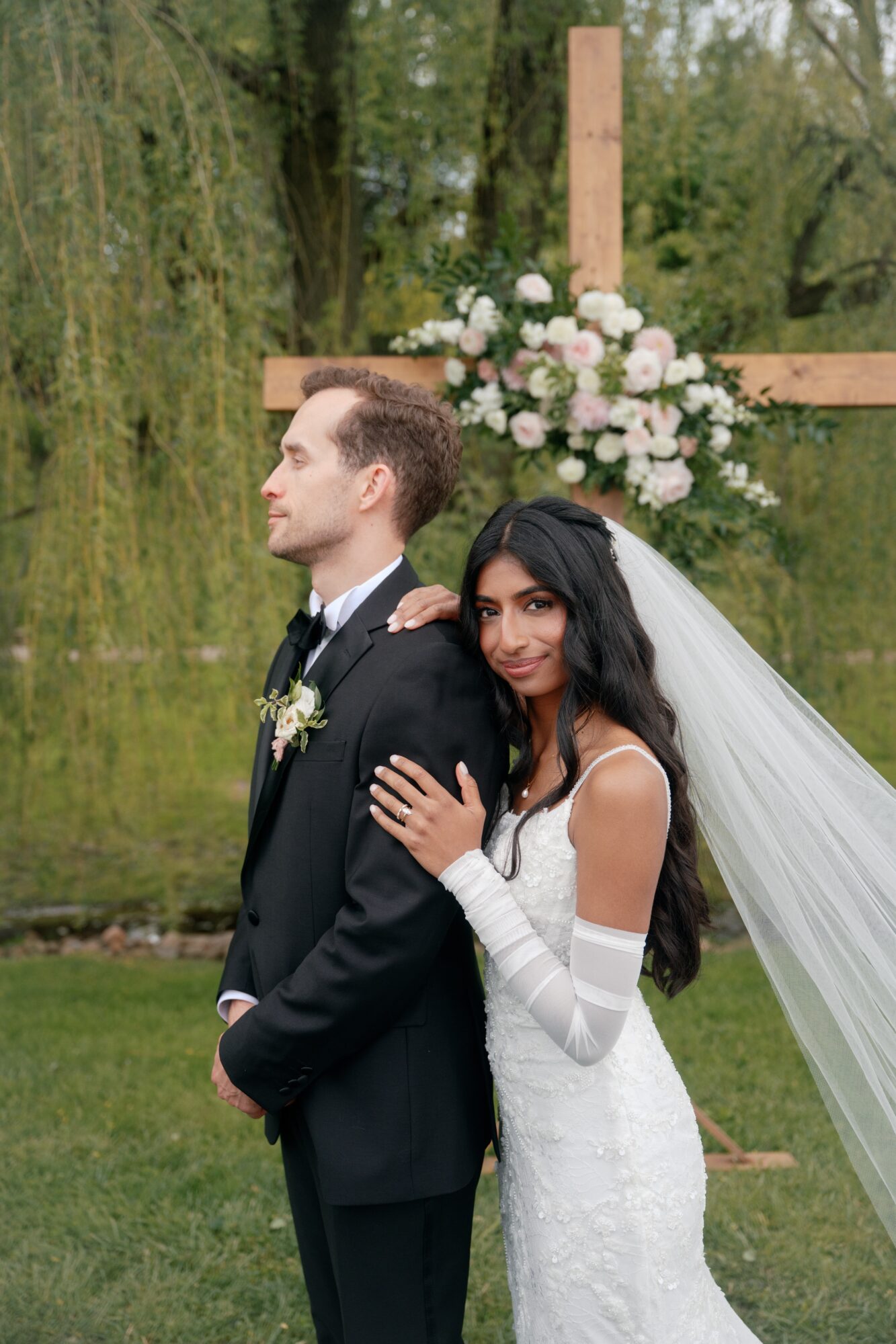 Bride and groom outdoors near a wooden cross with flowers, bride smiling, groom looking away, greenery background