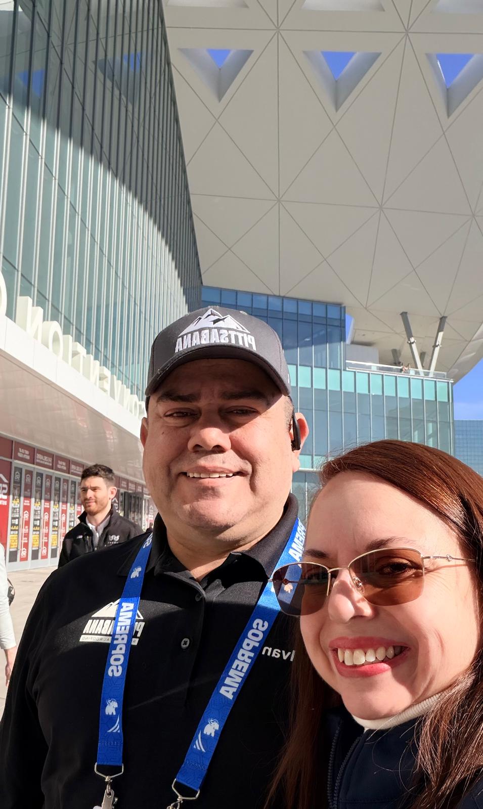 Two people smiling outdoors in front of a modern building with geometric architecture, one wearing a cap and the other sunglasses.