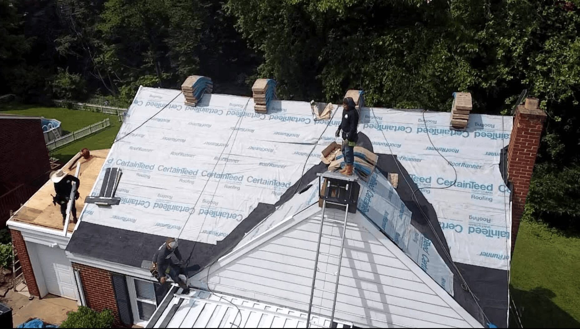 People working on a roof with a white underlayment, surrounded by trees and a grassy yard.
