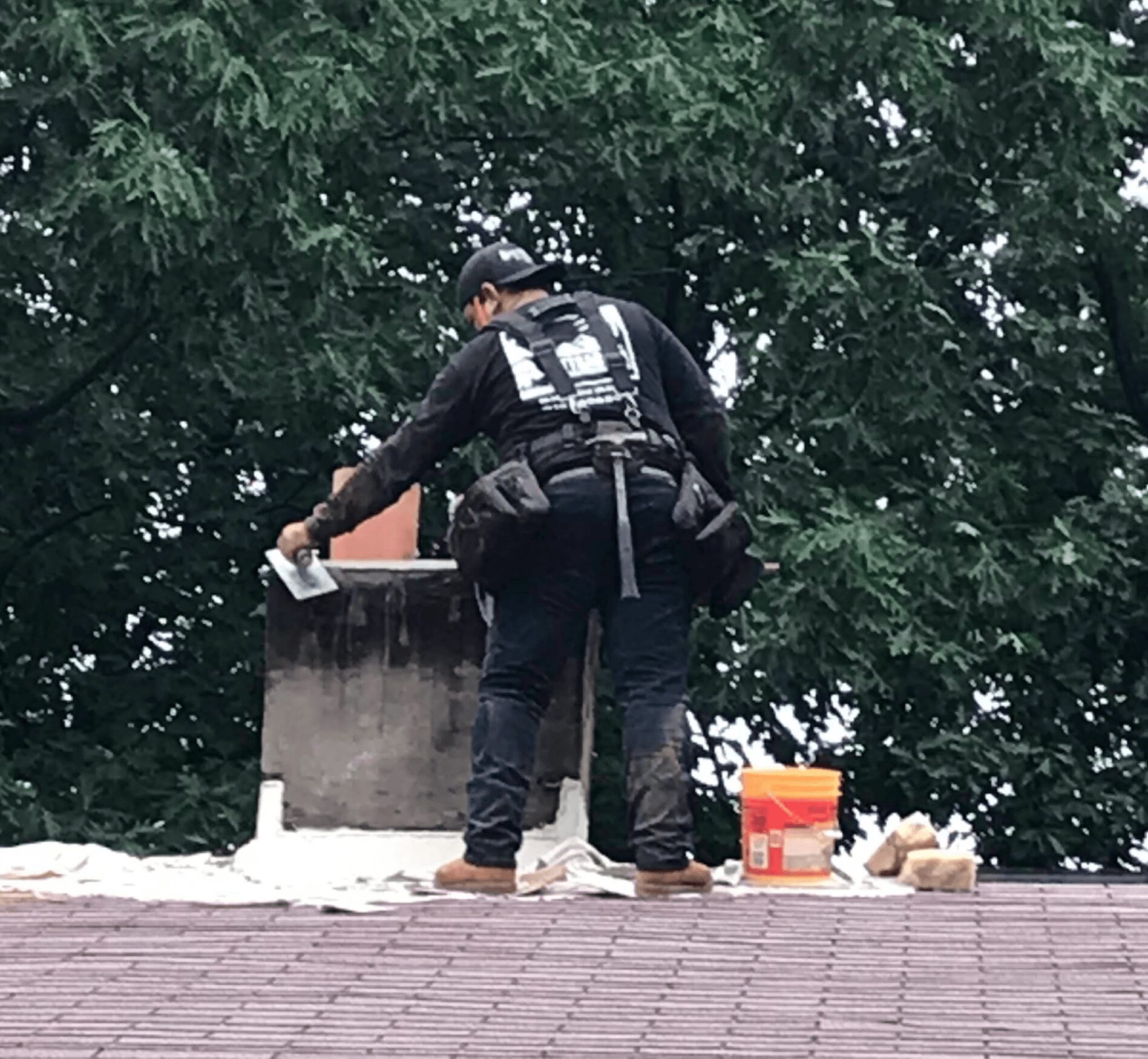 Person in black clothing and backpack working on a chimney or vent pipe on a roof, surrounded by trees and tools.