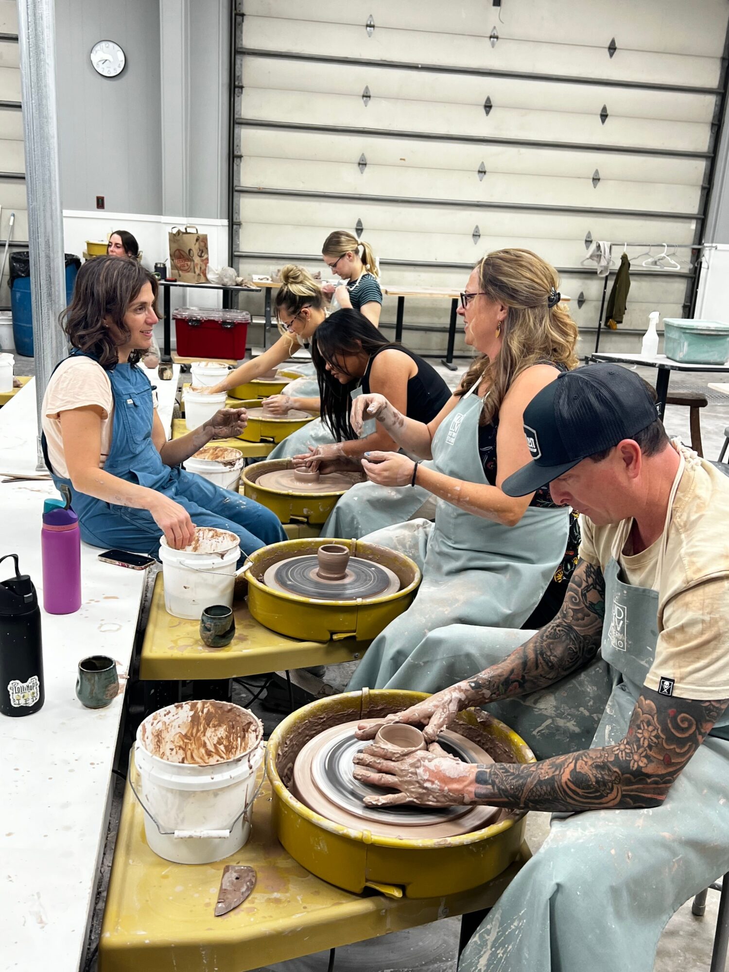 People working on pottery at a table inside a workshop, with a large garage door in the background.