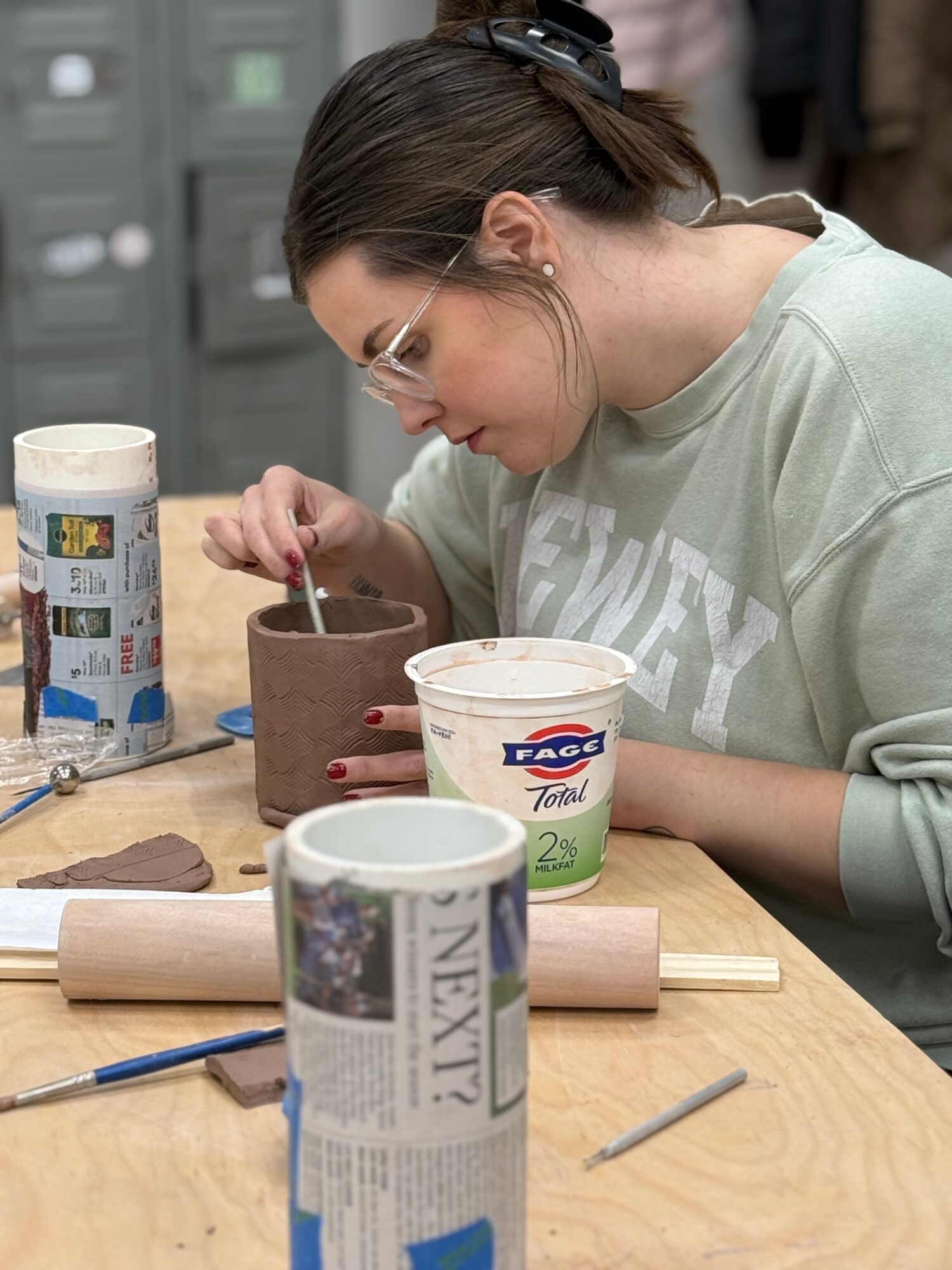 Young woman working on a pottery project at a table with clay and tools, wearing glasses and a light sweatshirt.