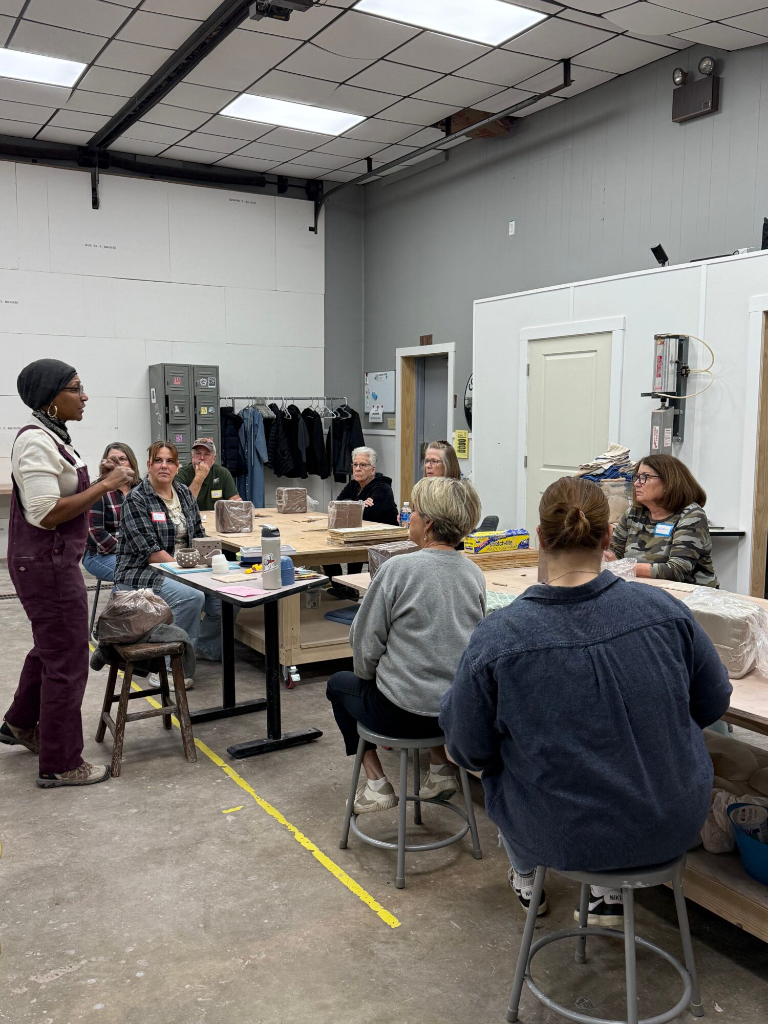People gathered around a table in a room with white walls and ceiling, some seated and some standing, engaging in discussion.