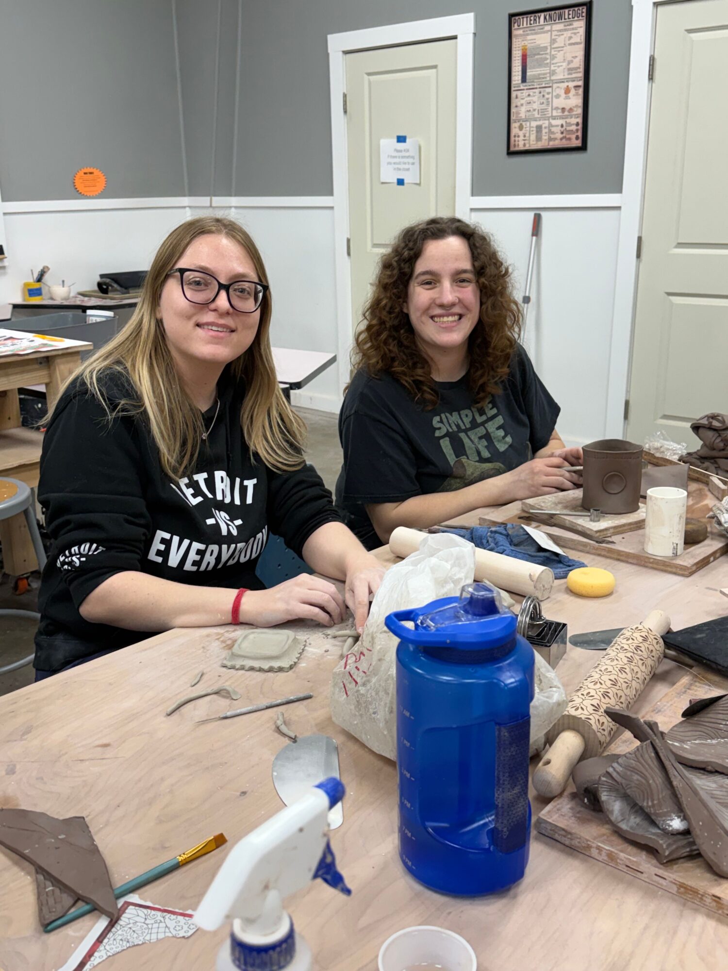 Two women sit at a table with craft supplies, smiling at the camera in a room with white cabinets and a door.