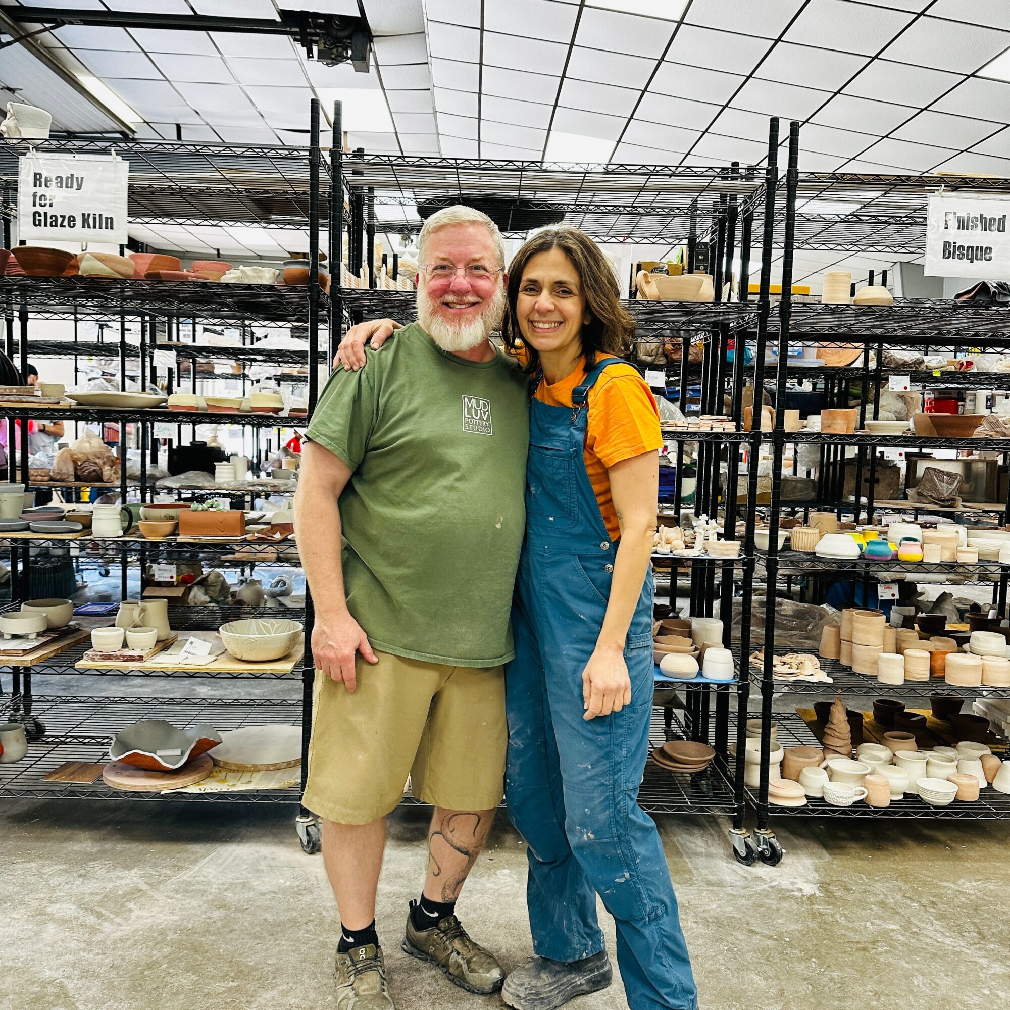 Two people, a man and a woman, stand together in a warehouse with shelves of pottery behind them, smiling.