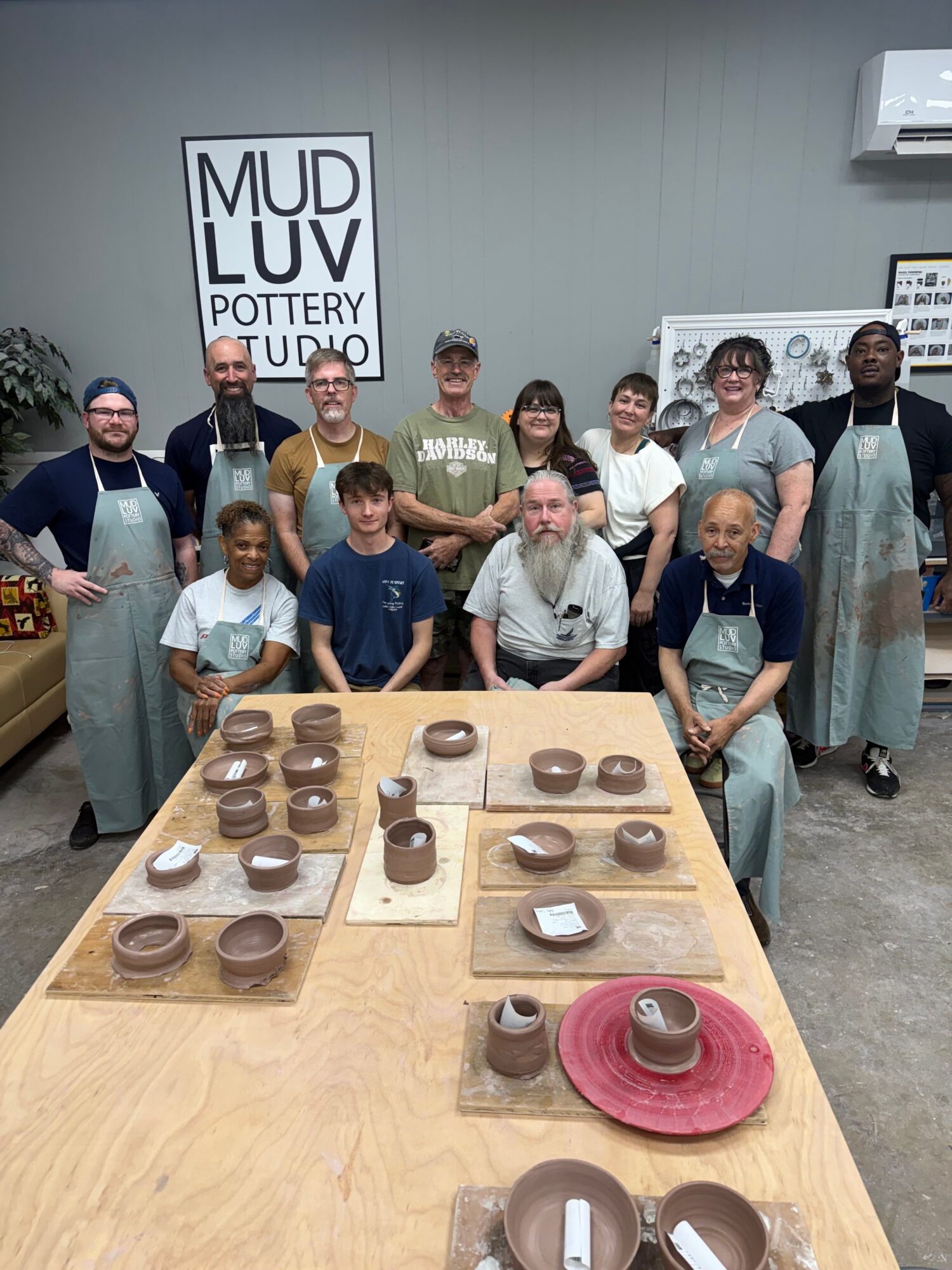Group of people standing behind a table with various small objects, in a room with a sign reading 'MUD LUV OUTPOST'.