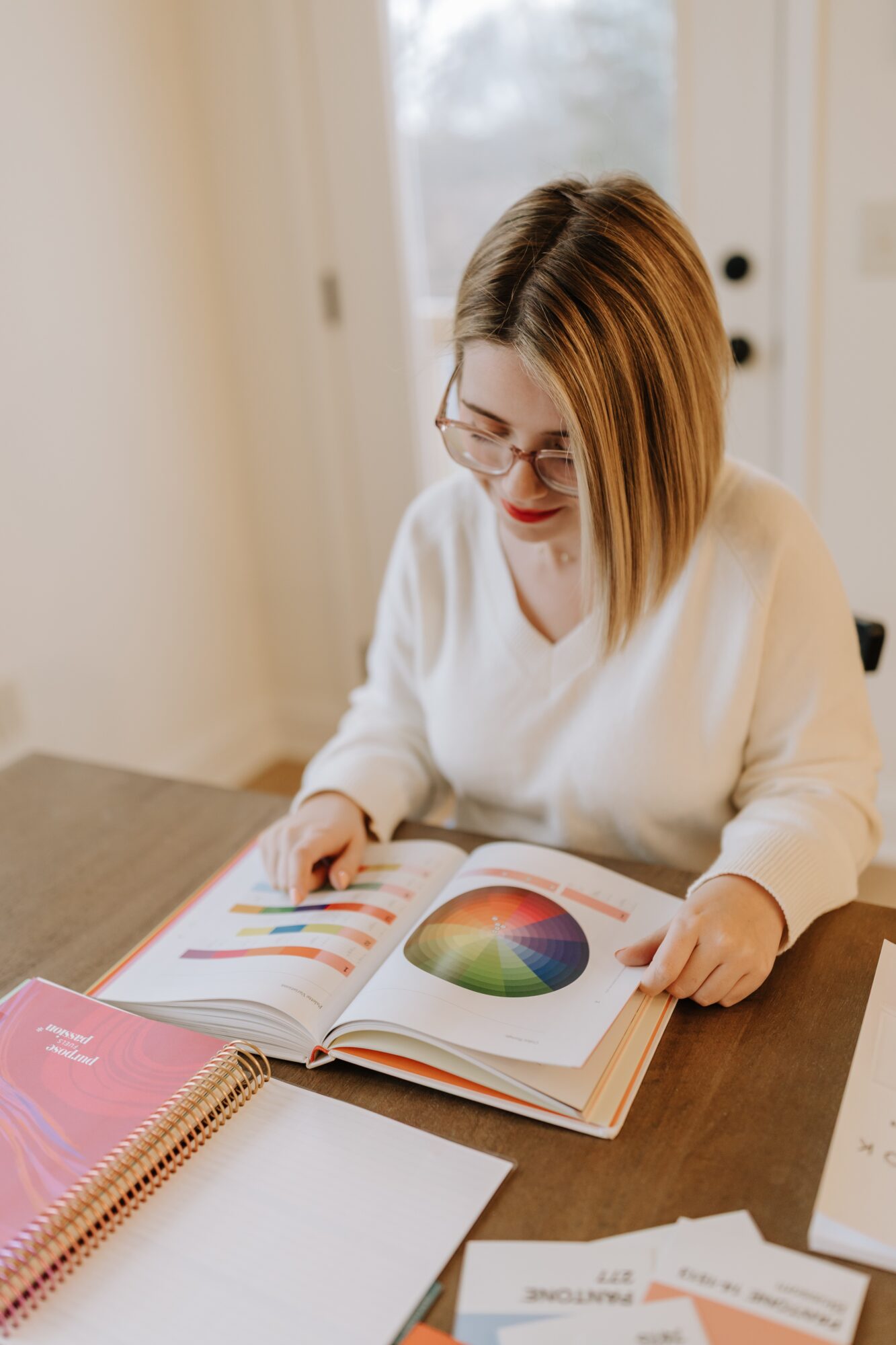 Woman with glasses and shoulder-length hair reads a colorful book at a table, smiling, in a bright room.
