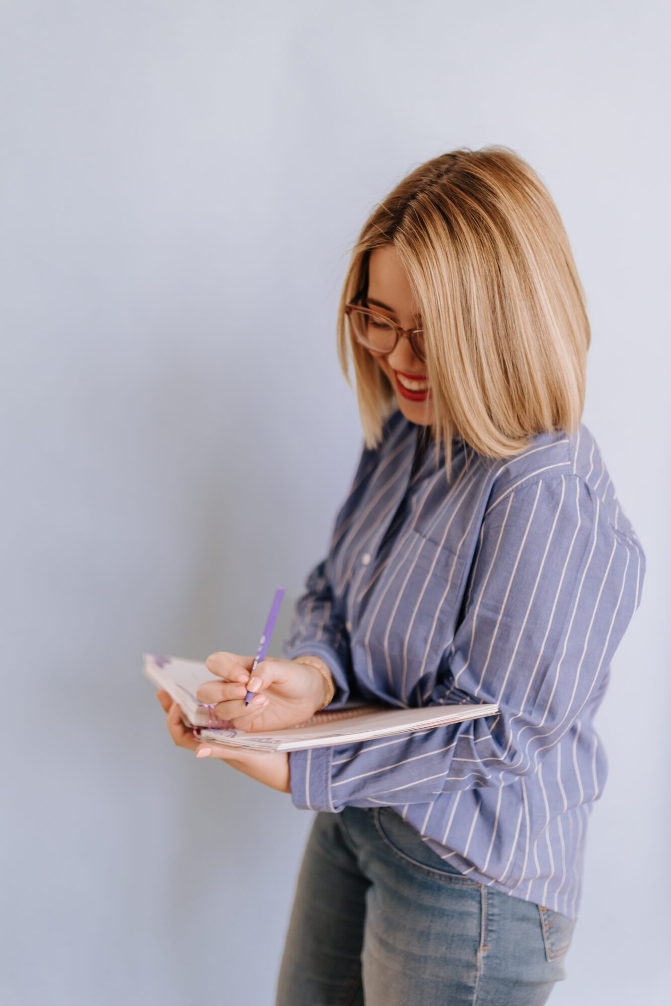 Woman with blonde hair and glasses writing in a notebook, wearing a striped shirt, smiling, against a light background.