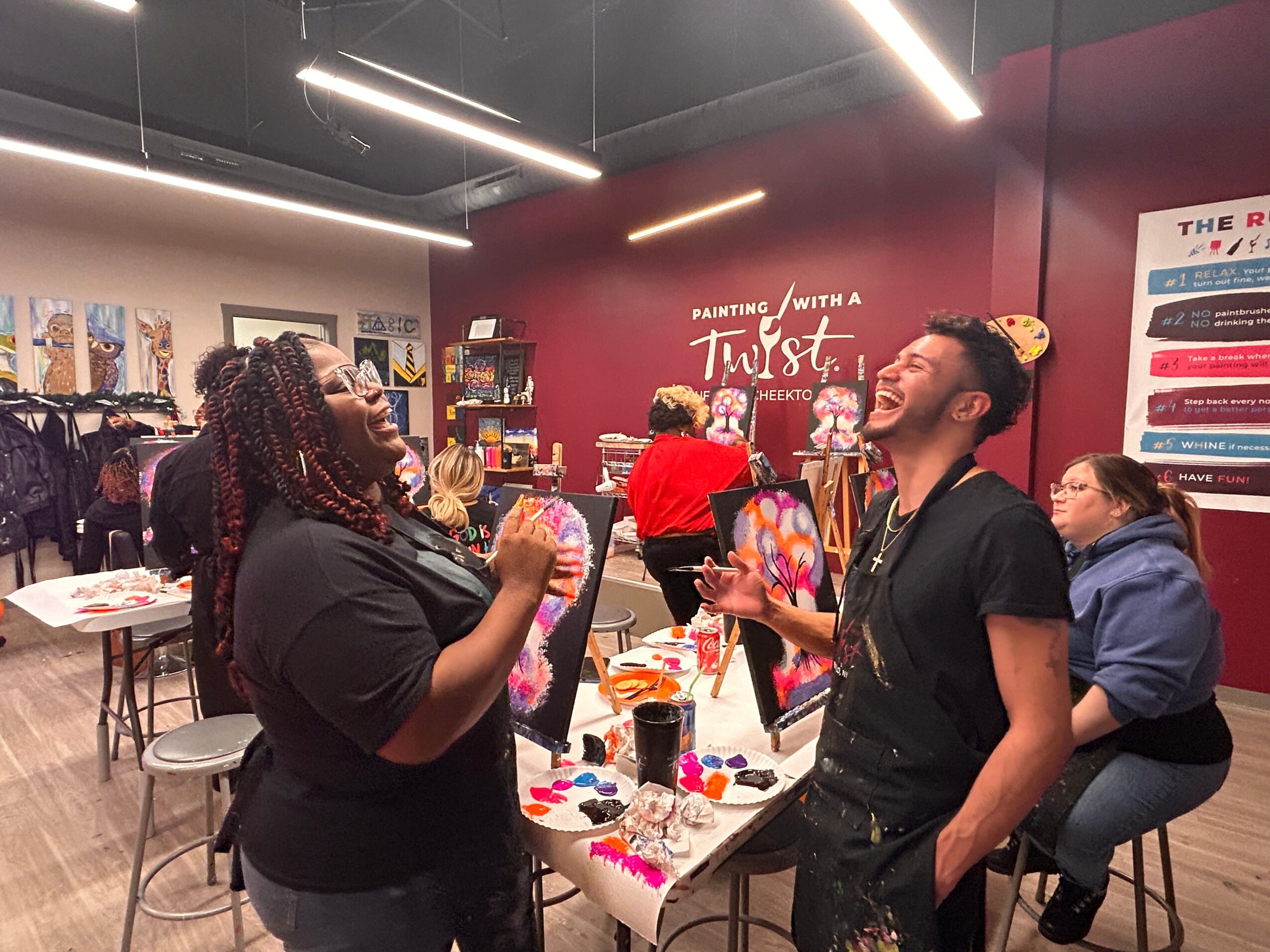 People participating in a painting activity at a store, with colorful paintings and art supplies on tables, and a red wall with text.