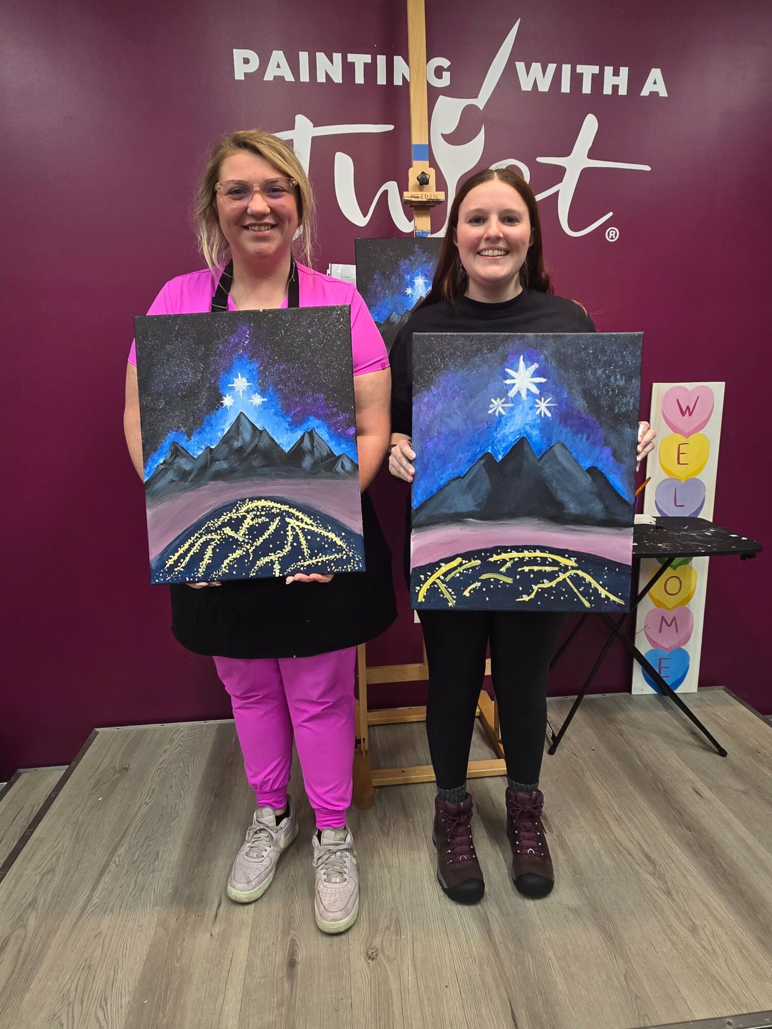 Two women holding paintings of mountains and a starry sky, standing in front of a purple wall with text, smiling.