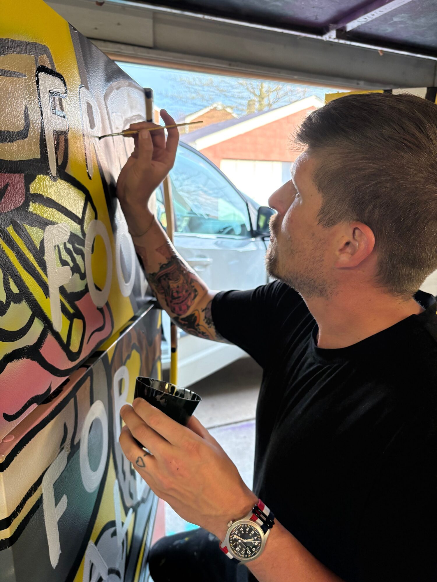 Person with tattoos painting a colorful mural on a large surface outdoors, with a car and building in the background.