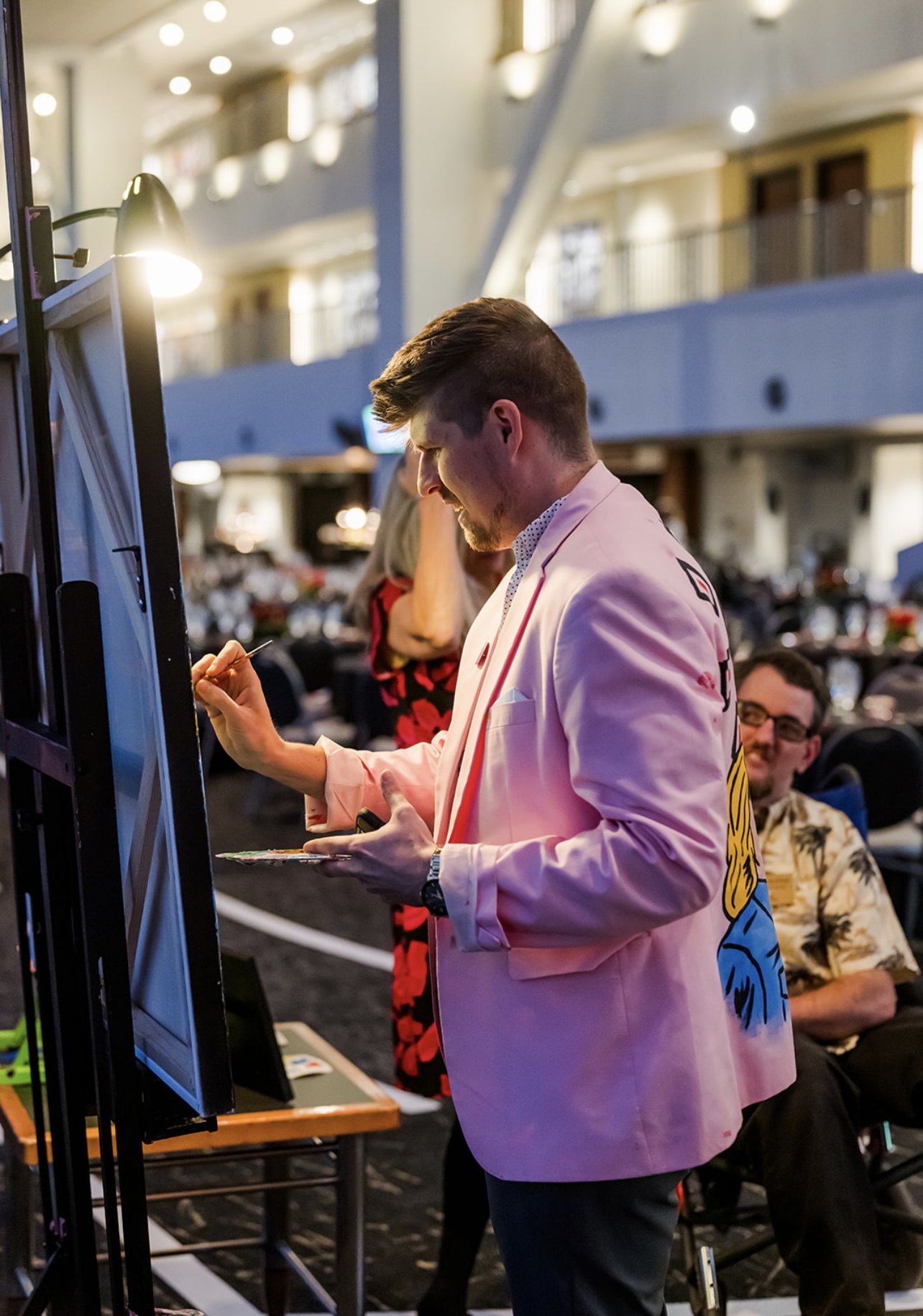 Person in pink blazer standing in front of a screen at an indoor event, with people seated in the background.