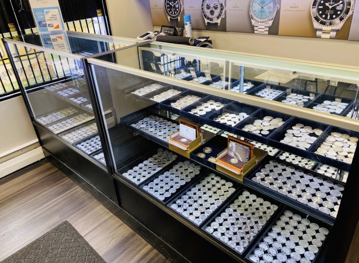 Display case filled with watches, with additional watches on the wall behind. Wooden flooring and a gray mat are visible.