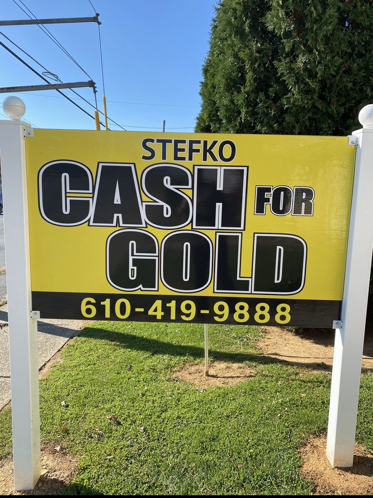 Sign advertising cash for gold services with phone number, yellow background, black and white text, outdoors with grass and trees