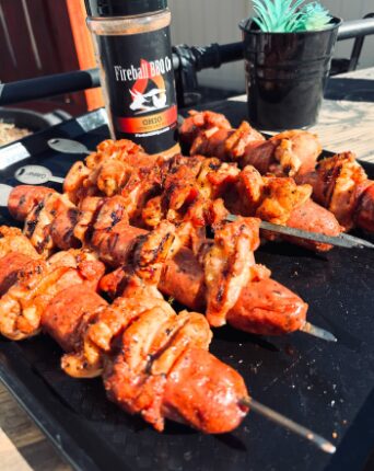 Skewers of grilled meat on a black surface with a bottle, a potted plant, and a bicycle handlebar in the background.