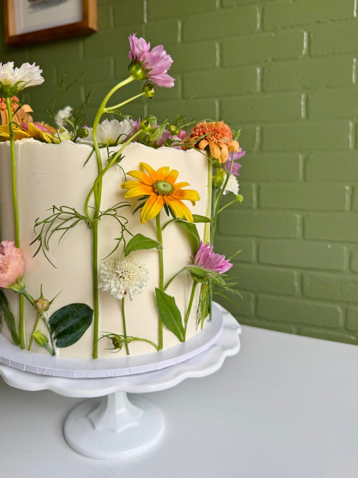 Cake decorated with colorful flowers against a green brick wall, on a white cake stand.