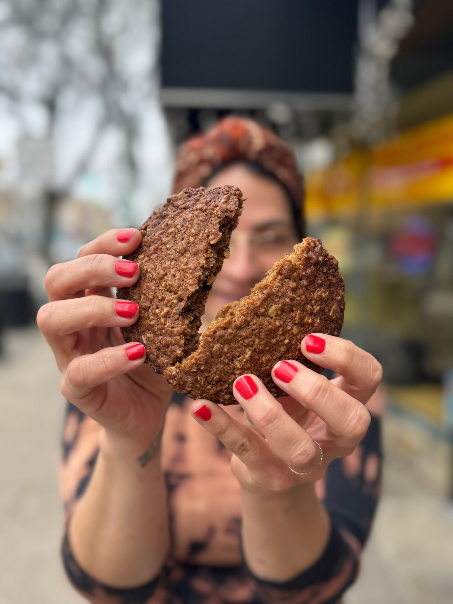 Person holding two large, breaded fried food pieces outdoors, with blurred background, red nail polish, and glasses.