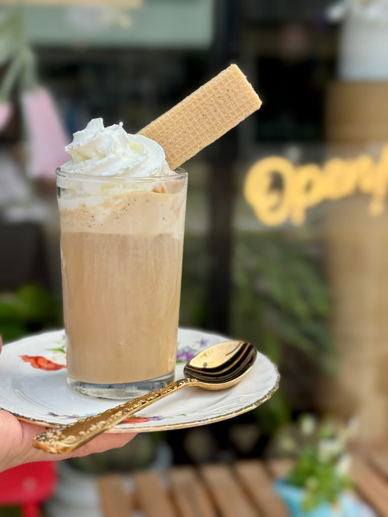 A glass of iced coffee topped with whipped cream and a wafer, on a decorative plate with a spoon, outdoors.