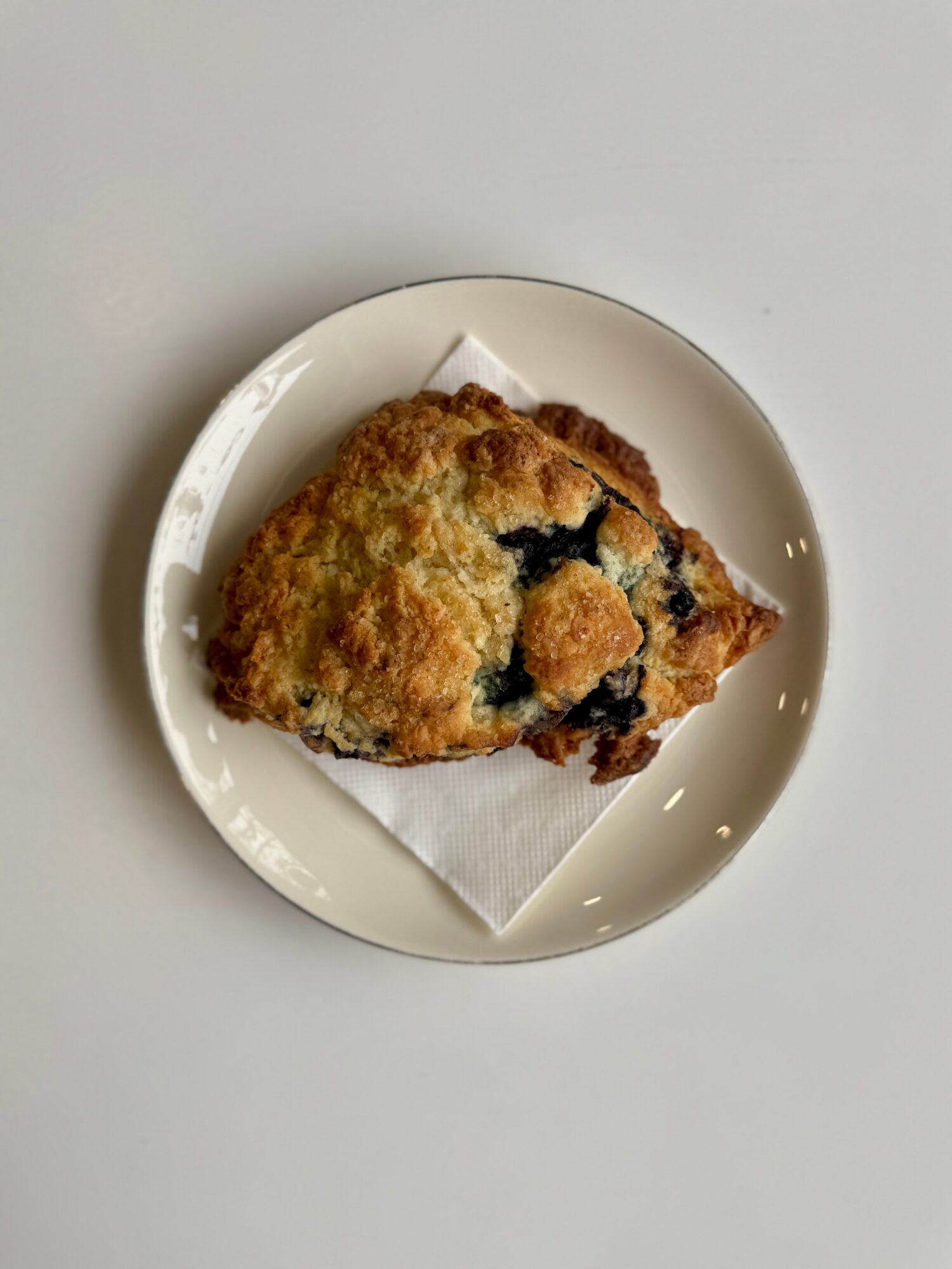 A slice of blueberry muffin on a white plate with a paper napkin underneath, placed on a white surface.