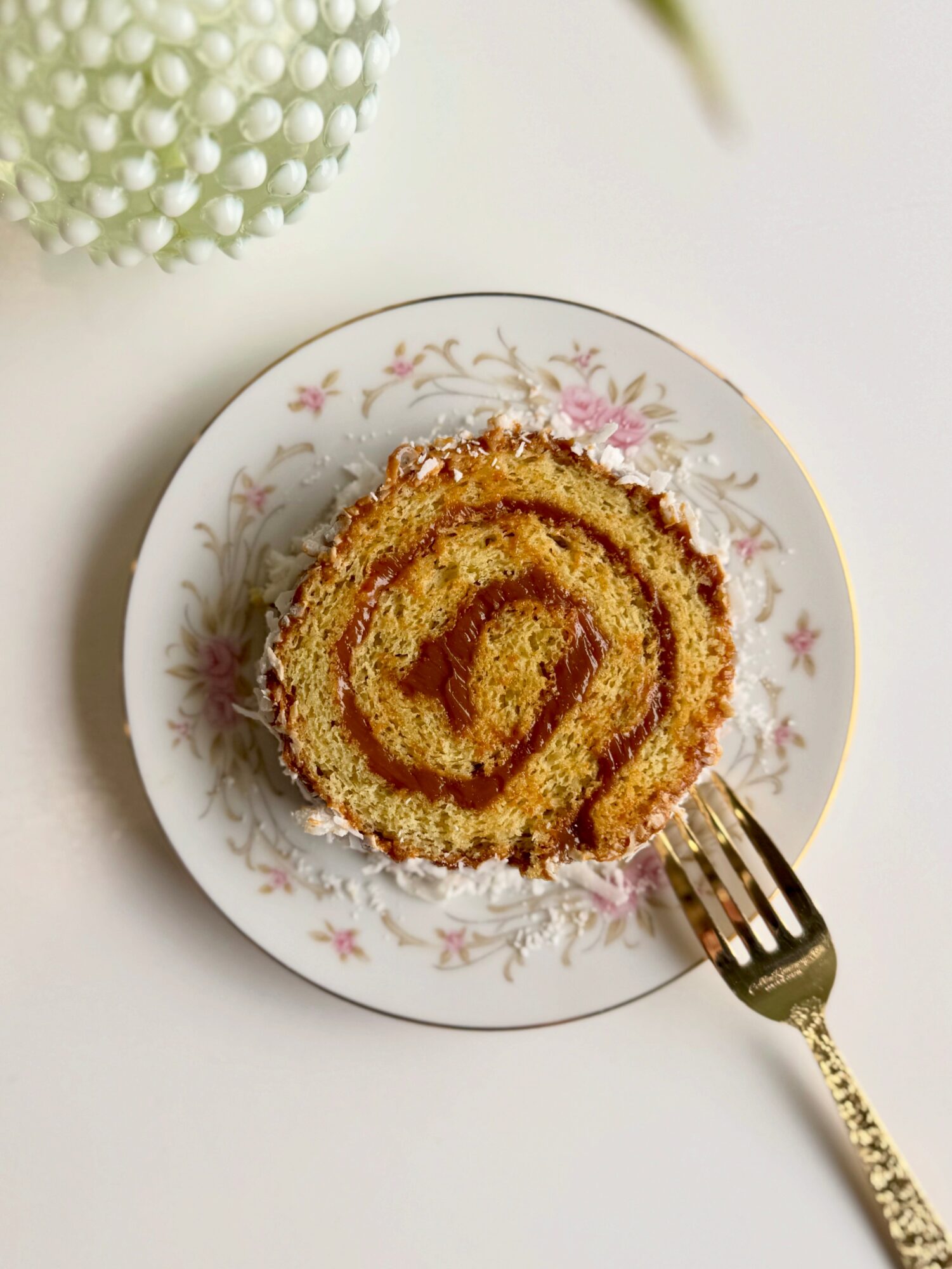 A slice of Swiss roll cake on a floral-patterned plate with a fork beside it, against a white background.