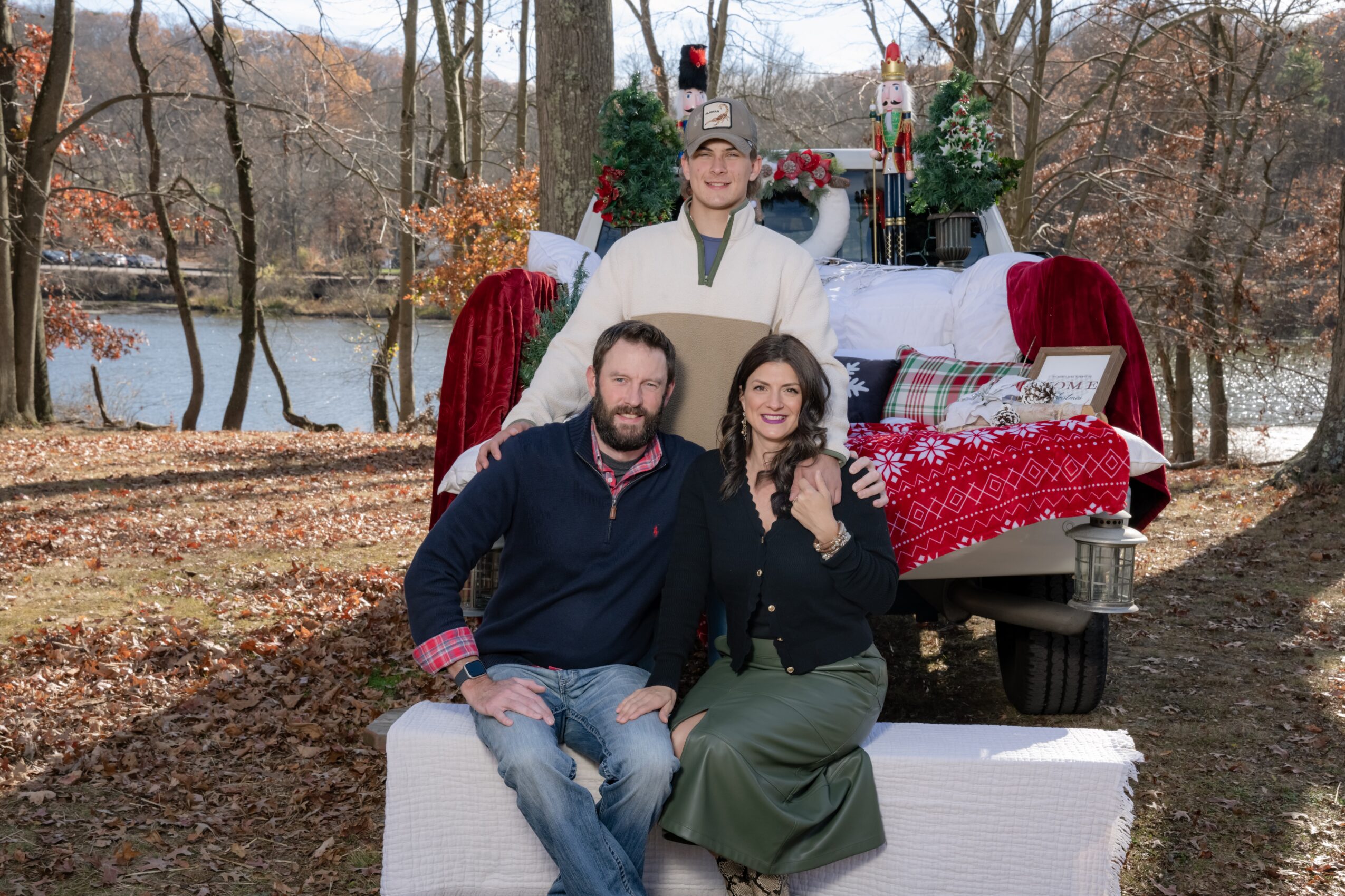 Three people outdoors near a decorated vehicle with a lake and trees in the background, autumn leaves on the ground.