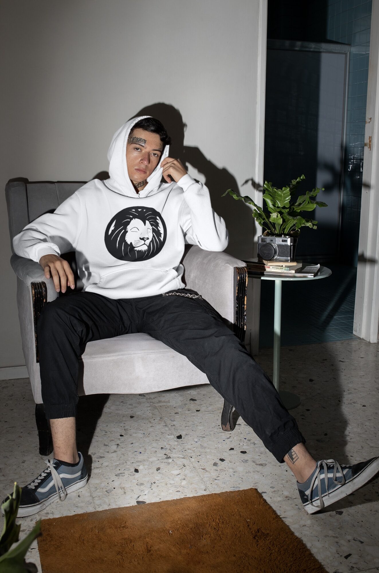 Person sitting on a chair indoors, wearing a hoodie and black pants, with a plant and window behind.