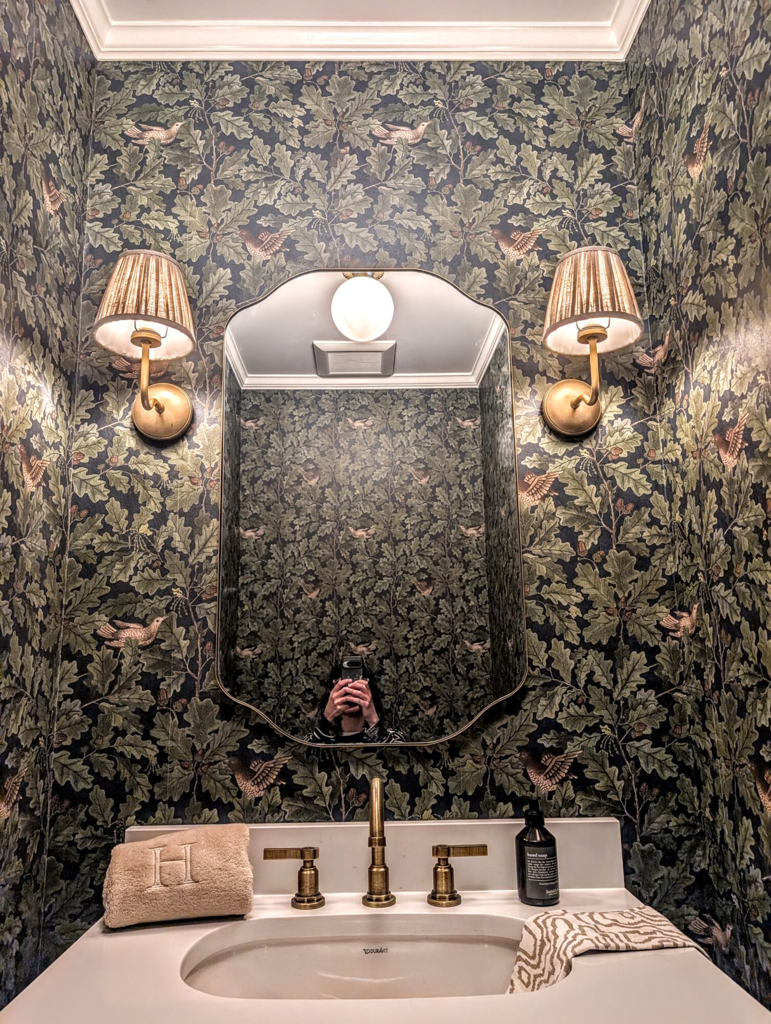 Decorative bathroom mirror with wall-mounted lamps, floral wallpaper, and a sink with soap and towel, viewed from below.
