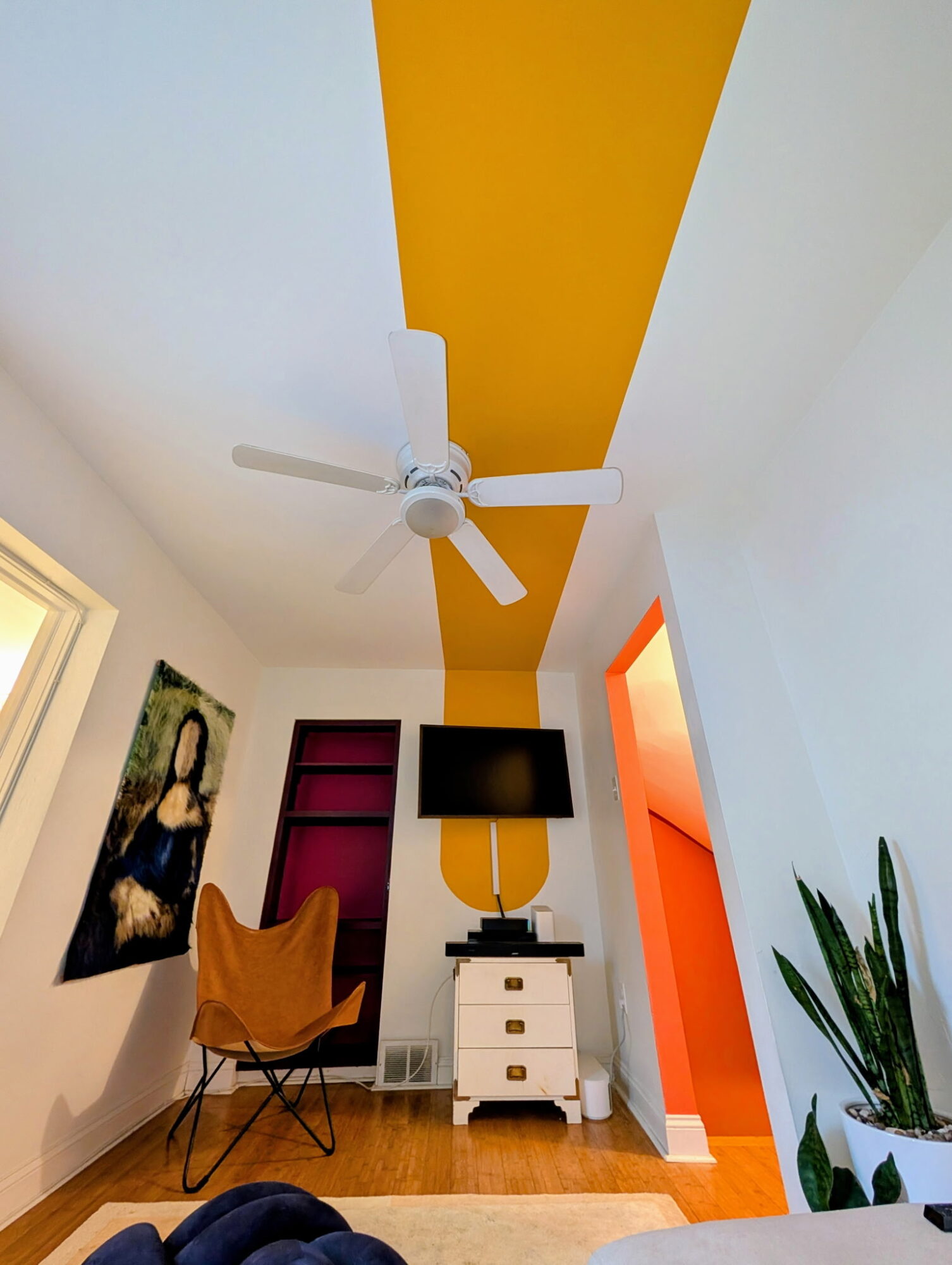 Room with a ceiling fan, yellow stripe on ceiling, TV on a white stand, orange doorway, and a plant.