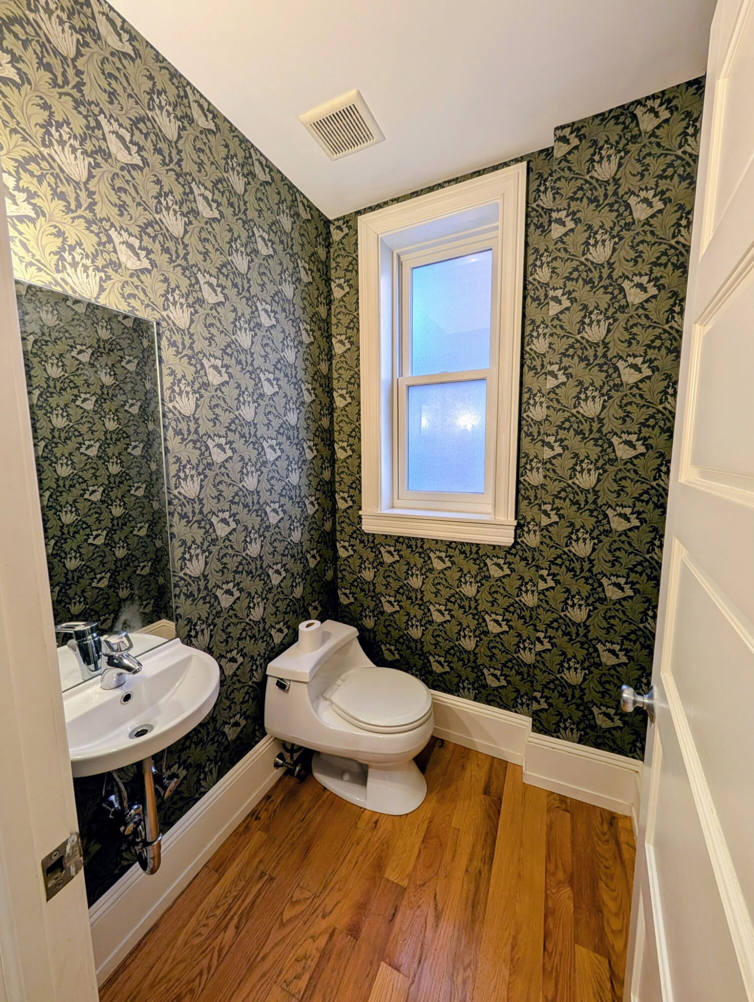 Small bathroom with floral wallpaper, white window, toilet, and corner sink, wooden floor, and white door partially open.