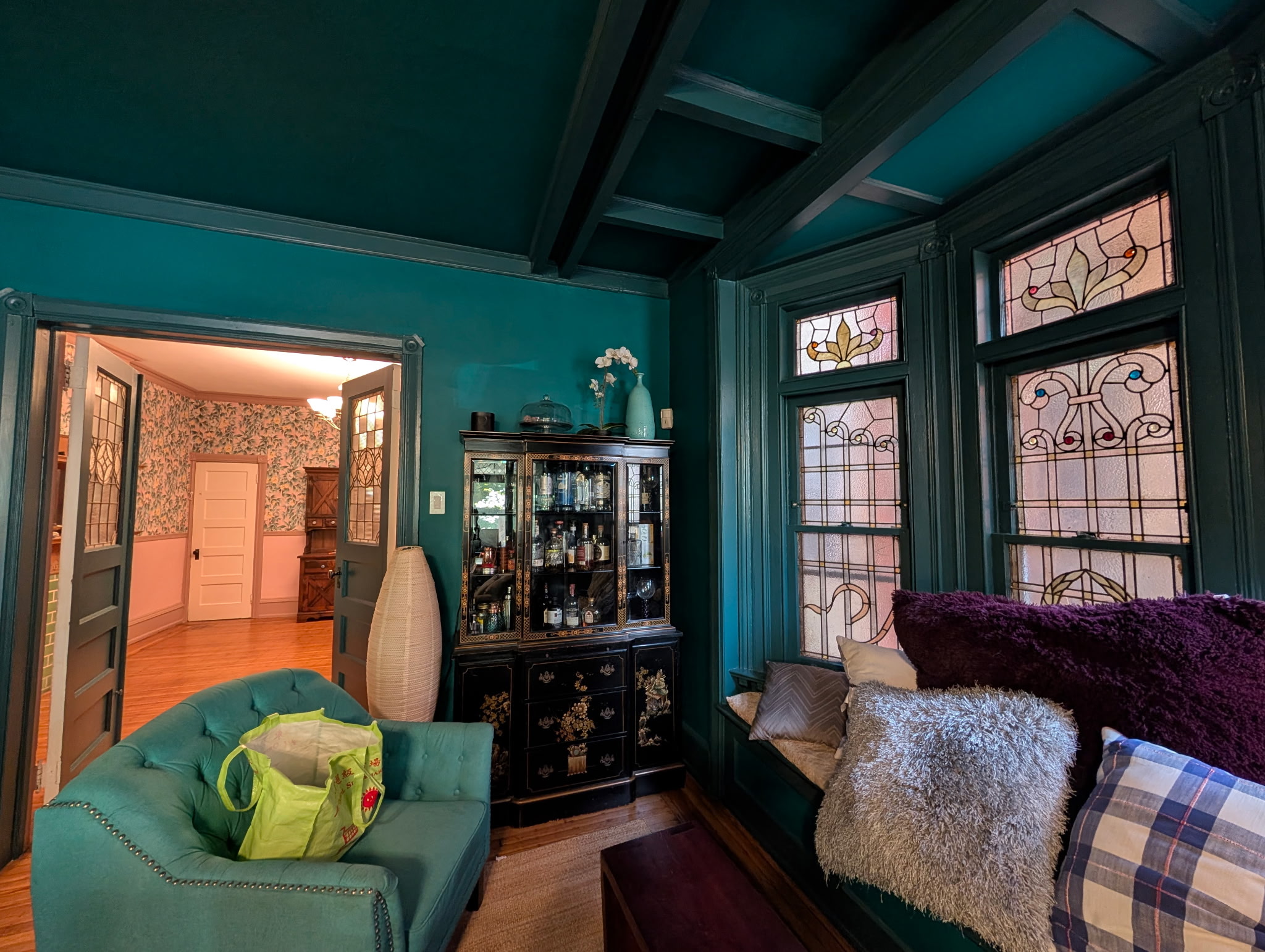 Living room with teal walls, large windows, a sofa with pillows, and a cabinet with glassware.