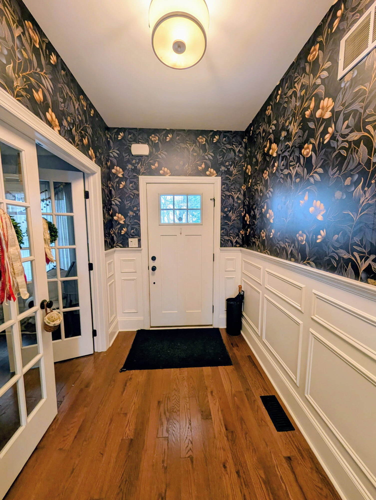 Hallway with floral wallpaper, white wainscoting, front door, and wooden floor, viewed from inside the house.