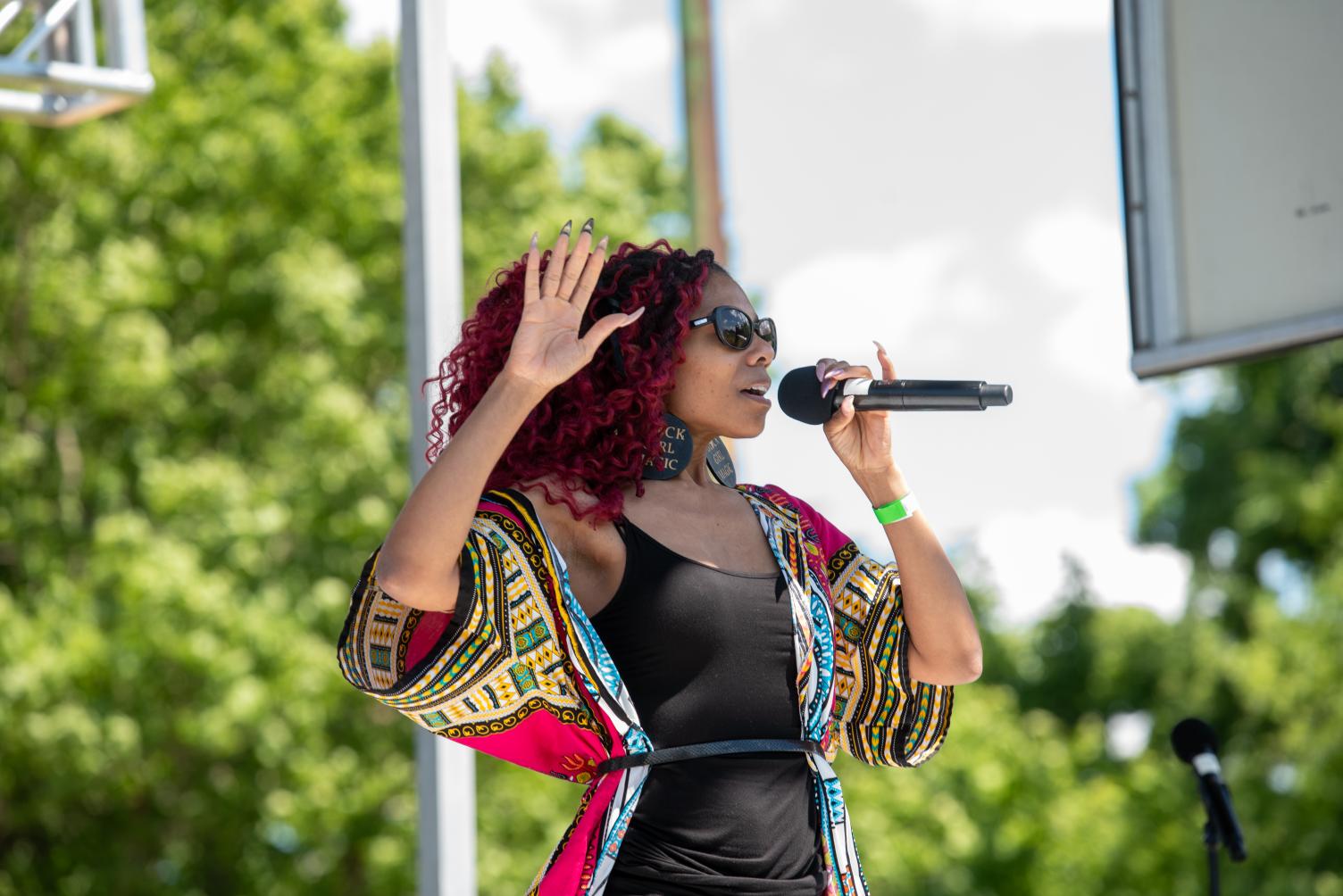 Woman with curly red hair wearing sunglasses and colorful jacket speaking into a microphone outdoors.