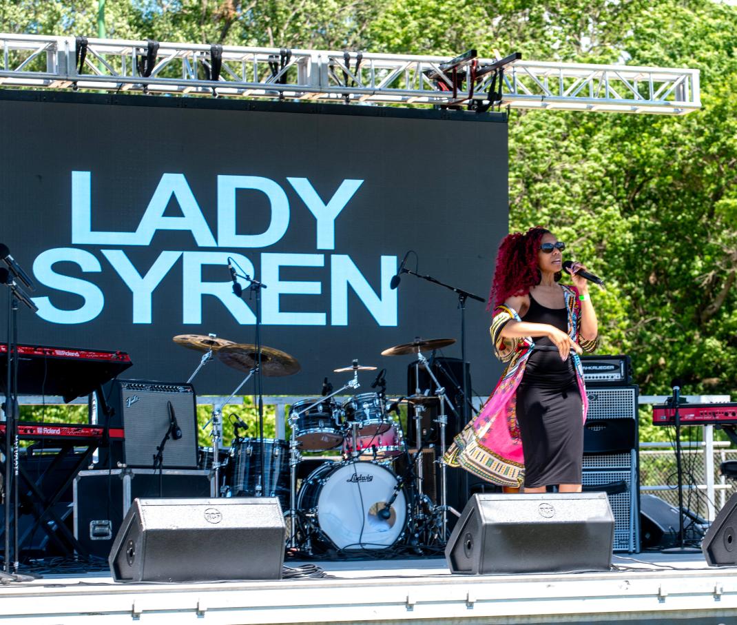 A woman with red hair and sunglasses sings into a microphone on an outdoor stage with musical instruments and a large screen behind her.
