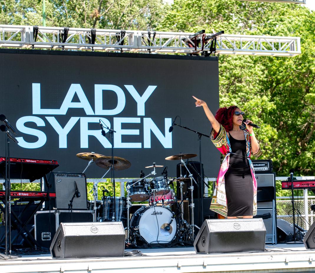 Performer singing on stage with a large screen behind displaying 'LADY SYREN', surrounded by musical instruments and equipment.