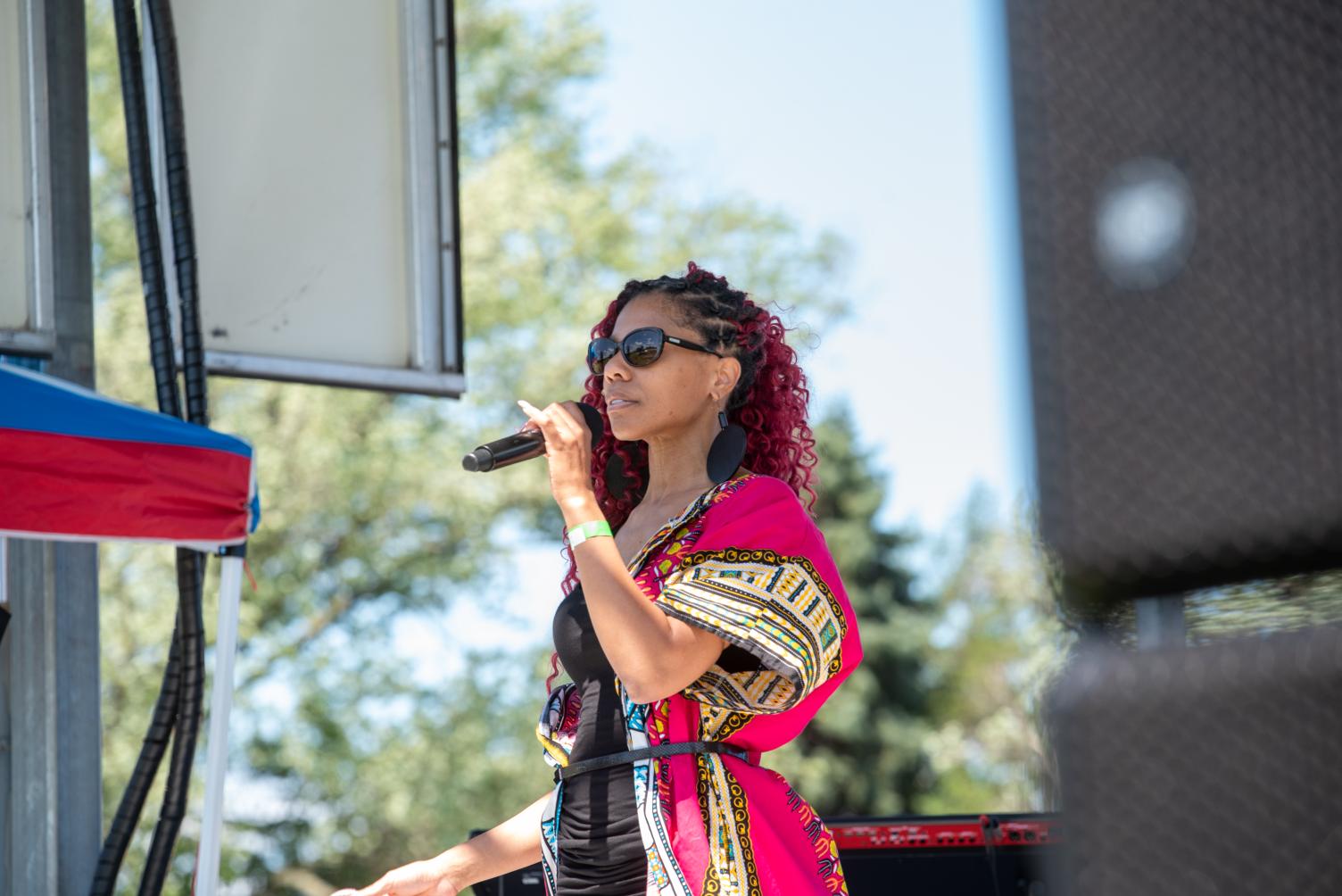 Woman with curly hair and sunglasses singing into a microphone outdoors, trees and a blue sky in background.