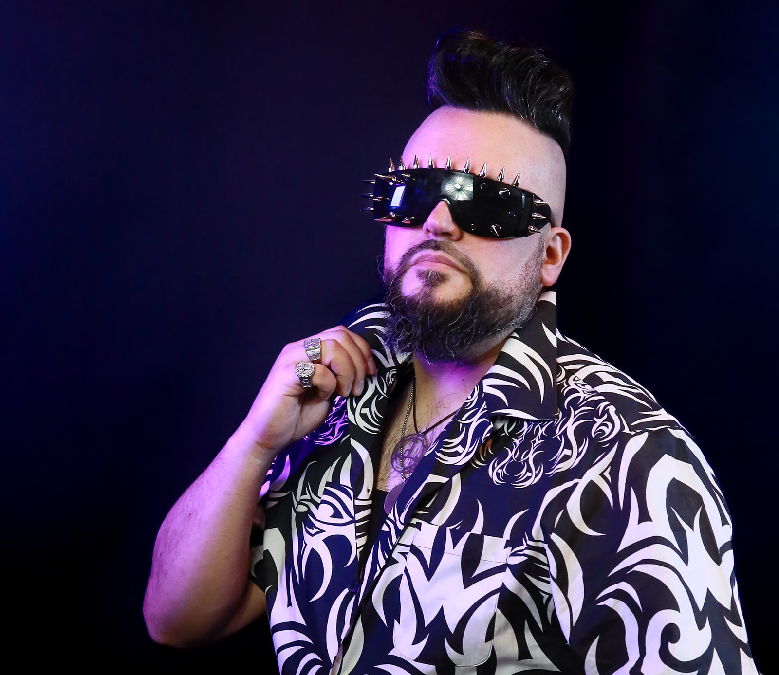 Person with styled hair, sunglasses, and patterned shirt, posing with hand near face against dark background.