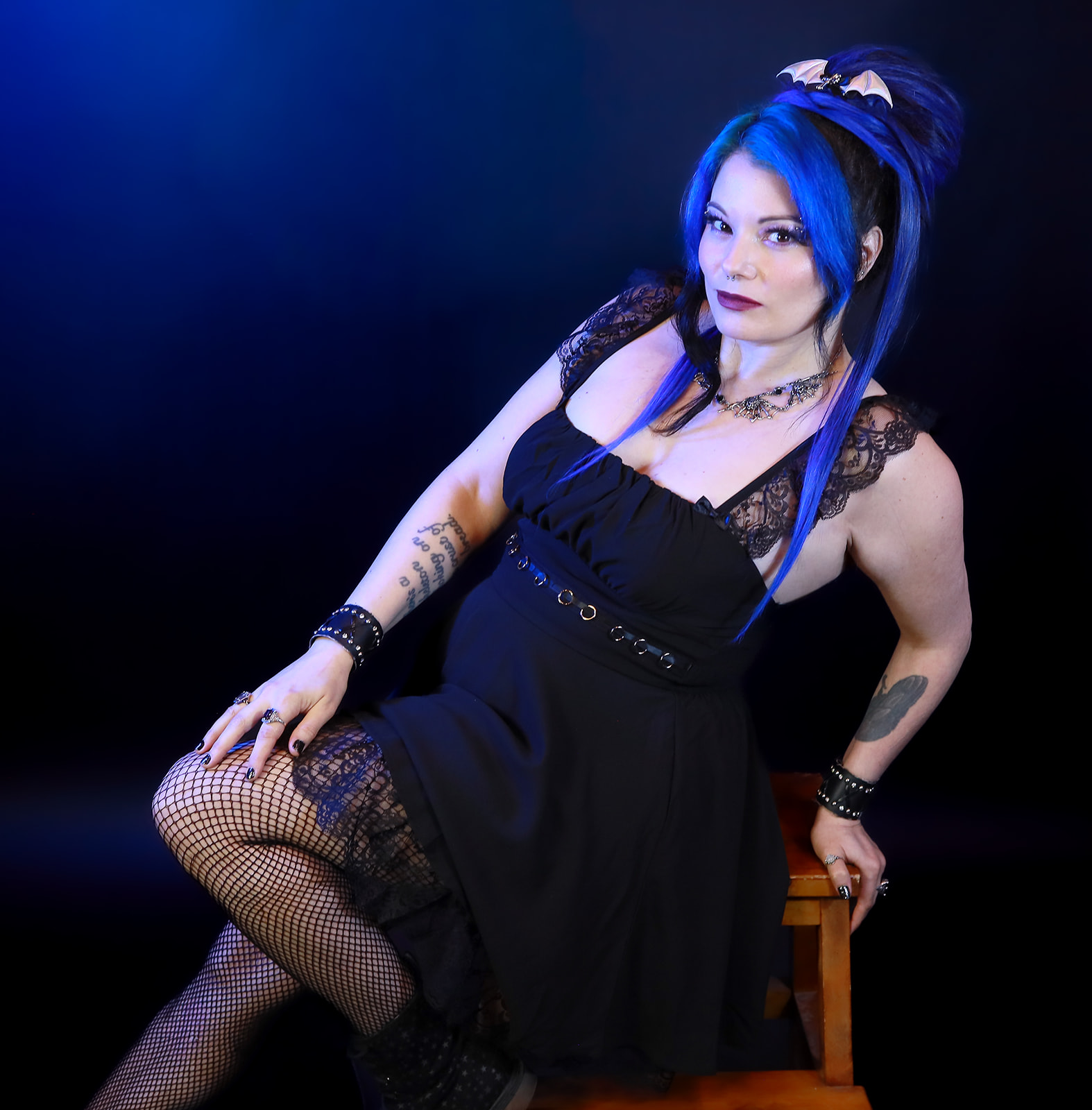 Woman with blue hair in a black dress, sitting on a chair, with dark background.