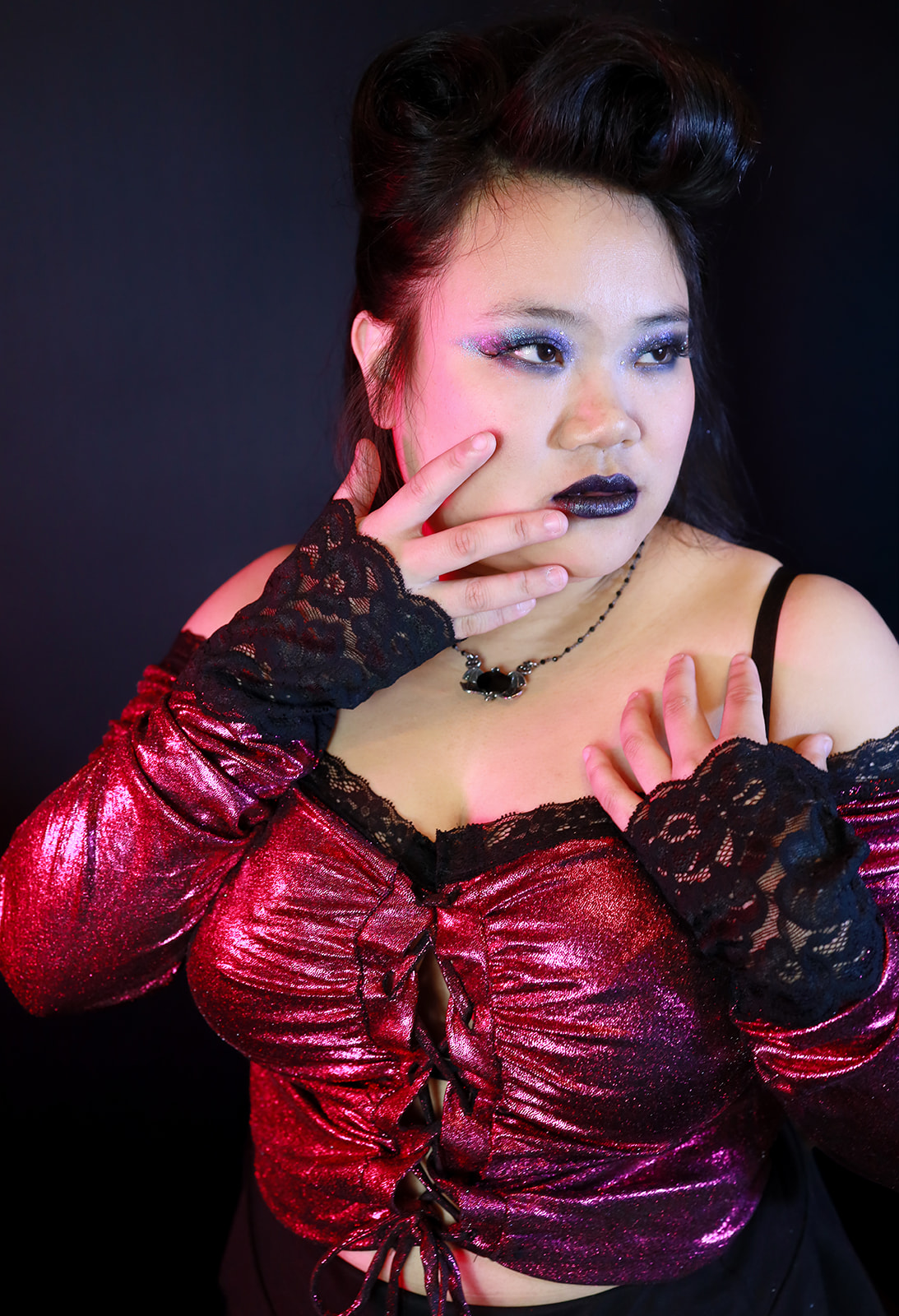 Woman with dark hair, makeup, and gothic attire, touching her face with gloved hands, against a dark background.