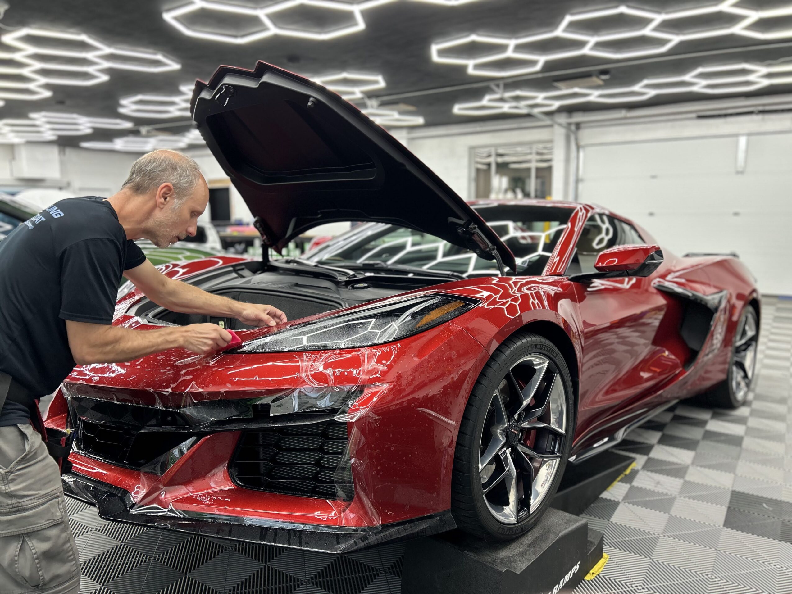 Man working on a red sports car with open hood in a garage, checkered floor, modern ceiling lights.