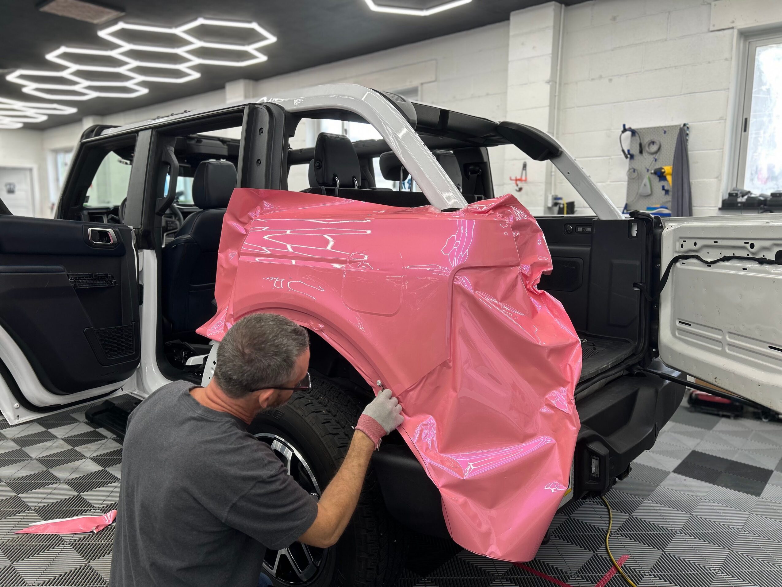 Person working on a pink vehicle fender inside a workshop with black and white checkered floor and modern ceiling lights.
