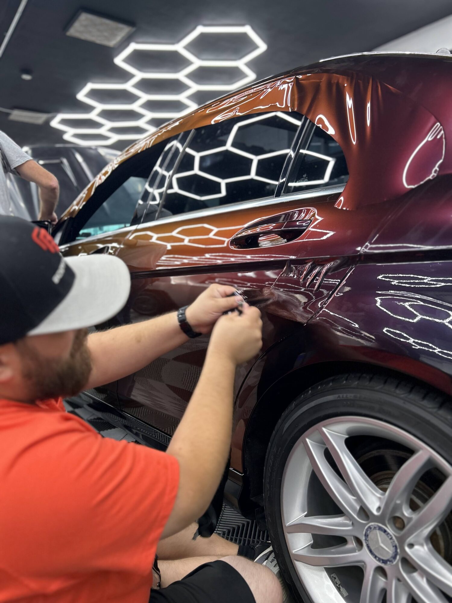 Person in red shirt and cap working on a shiny maroon car in an indoor showroom with hexagonal ceiling lights.