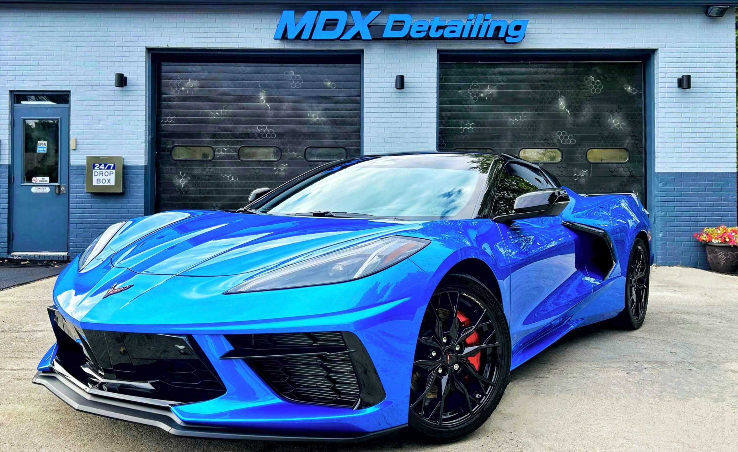 Blue sports car parked outside a detailing shop with closed garage doors and a sign reading MDX Detailing.