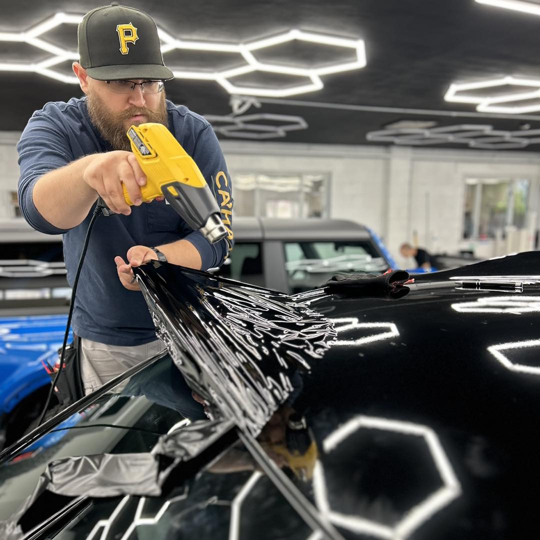 Man using a yellow heat gun to dry a black car in a modern, well-lit showroom.