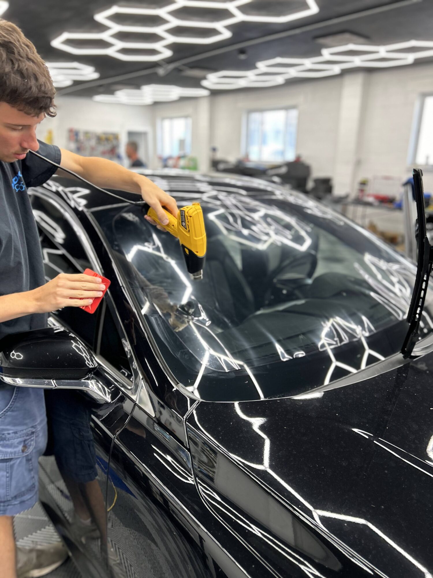 Person cleaning a black car with a yellow cloth in a well-lit garage, ceiling with geometric light fixtures, windows in background.